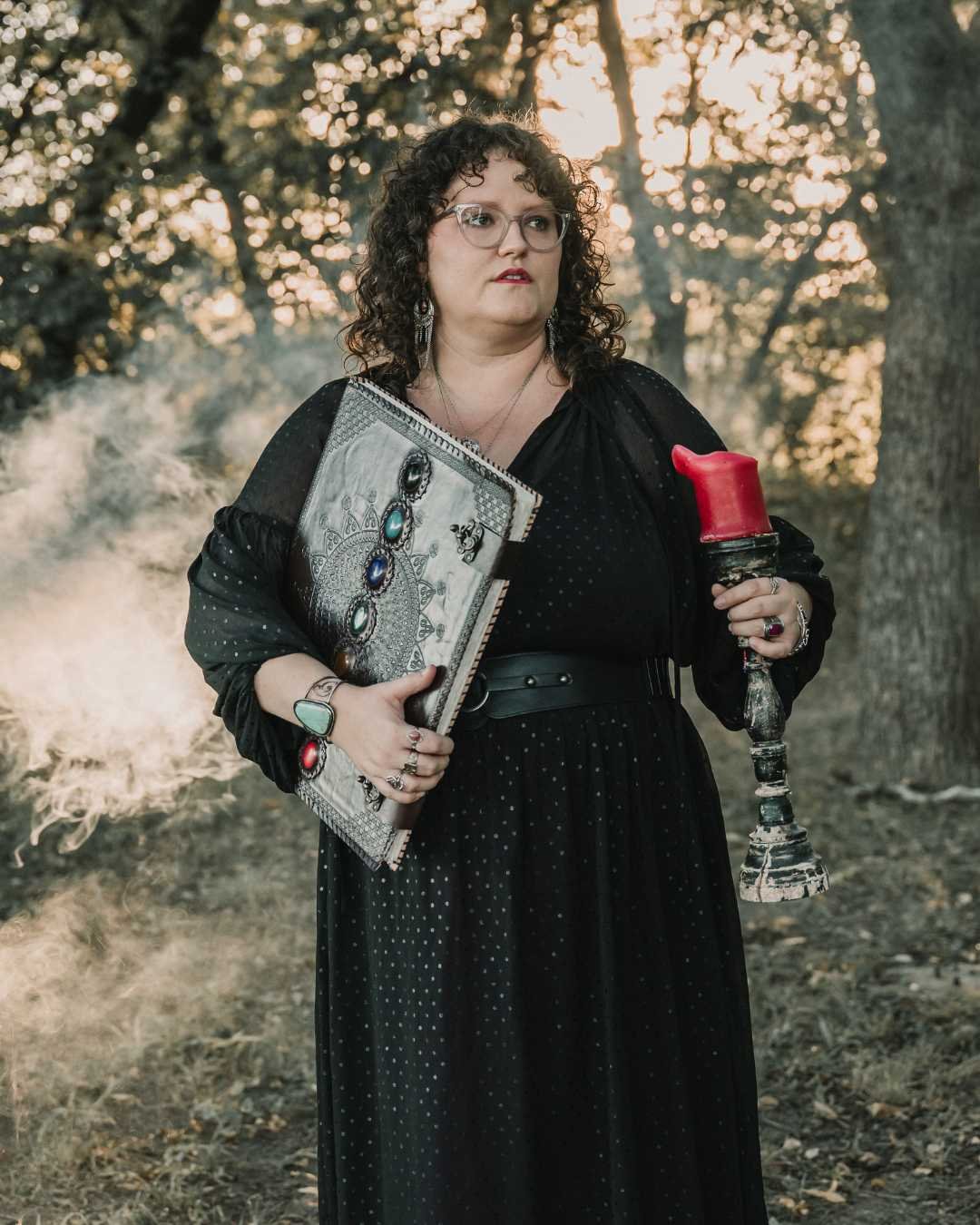 A woman dressed in black with glasses and curly hair holds a large decorated book or journal in one hand and a red candle in a vintage holder in the other, standing outdoors in a wooded area with sunlight filtering through the trees.