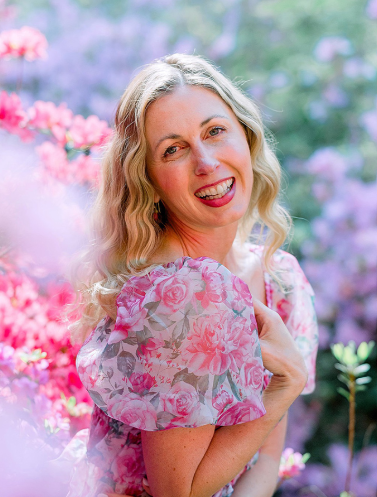 A woman with curly blonde hair smiling in a pink floral dress outdoors among blooming flowers.