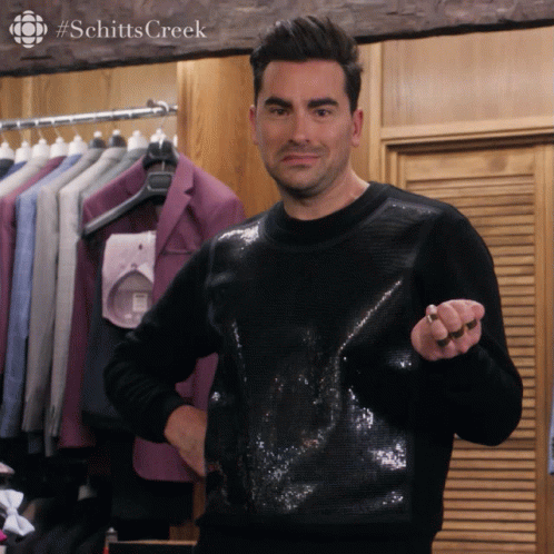 A man with dark hair and a slight smile is standing in a clothing store, holding a ring in his right hand. Behind him are suits and shirts hanging on a rack.
