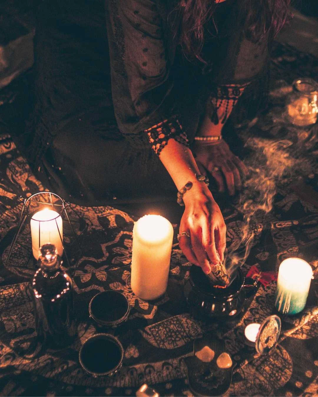 A woman practicing tarot reading in a dimly lit room with candles, bottles, and crystals on a patterned cloth.