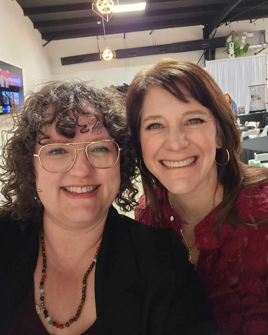 Two smiling women taking a selfie in an event space with tables and a television screen in the background.