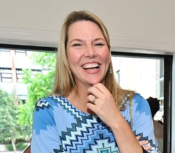 A woman with blonde hair smiling and touching her chin, wearing a blue patterned top, indoors with a window showing trees outside.