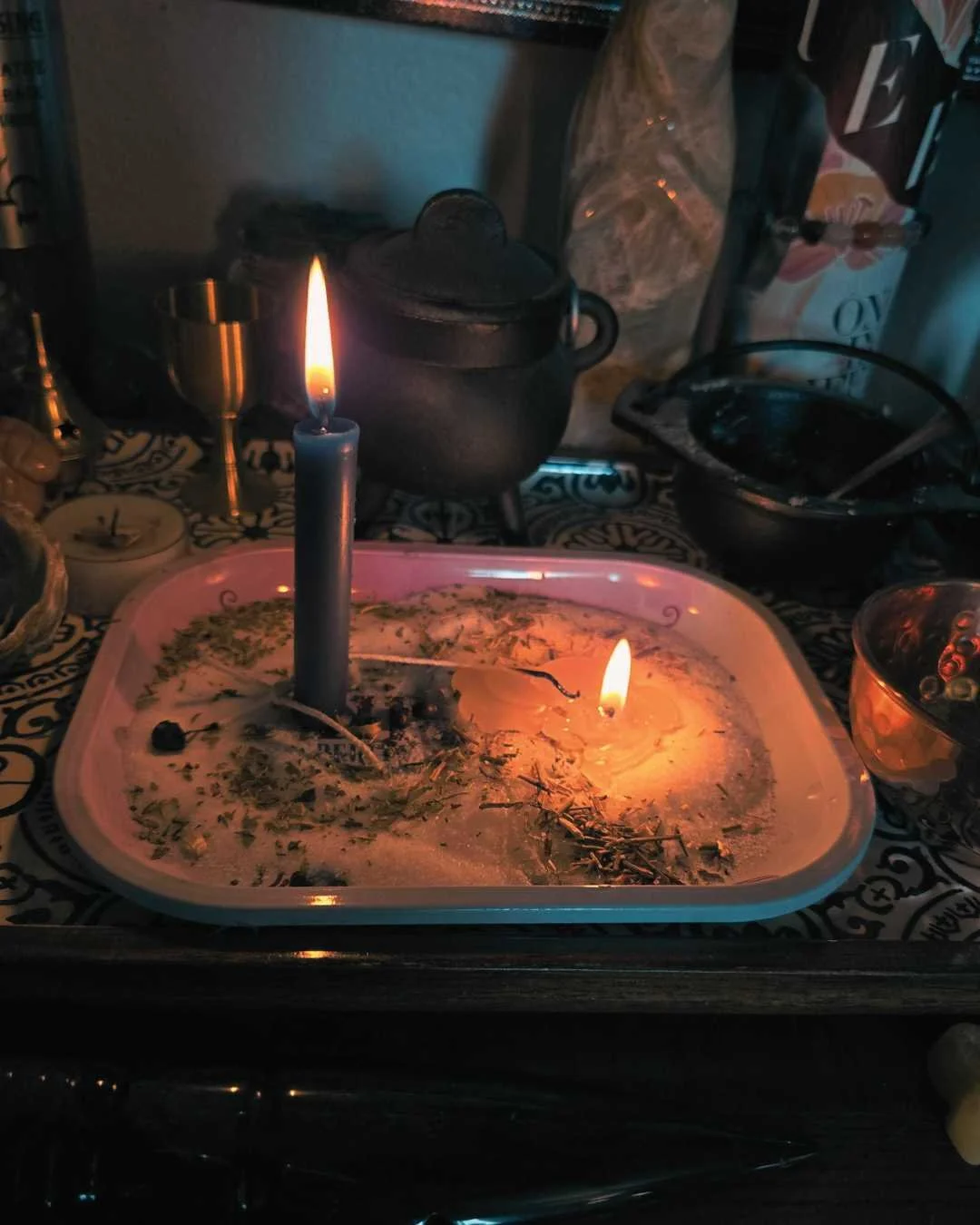 A lit candle on a dish with ashes, herbs, and small objects, surrounded by various kitchen items and utensils on a patterned table.