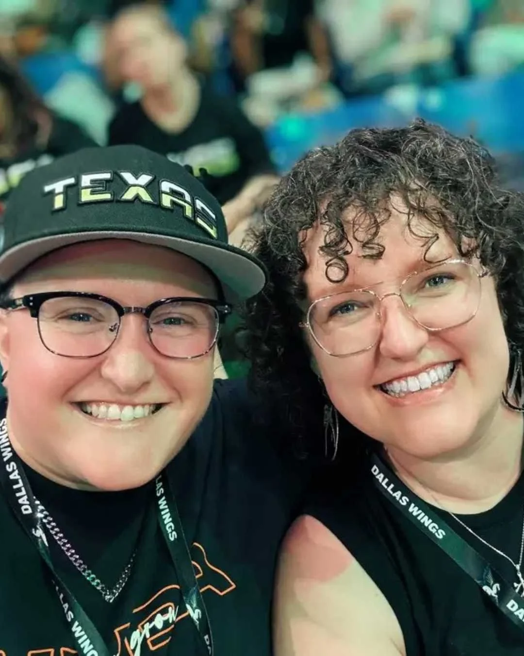 Two smiling women wearing glasses and black shirts, with one woman wearing a Dallas Wings lanyard and the other a Texas cap, taking a selfie at a sports event.