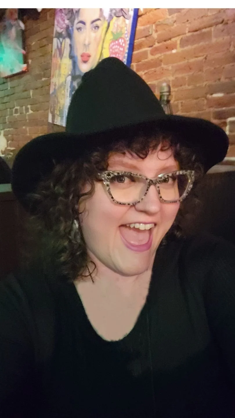 A woman with curly hair wearing leopard print glasses and a black wide-brimmed hat, smiling in a lively setting with a colorful painting of a woman's face on a brick wall in the background.