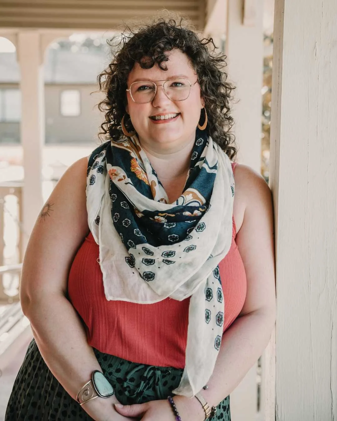 Smiling woman with curly hair, glasses, wearing a coral tank top, patterned skirt, and a navy and cream scarf, standing near a wooden post.