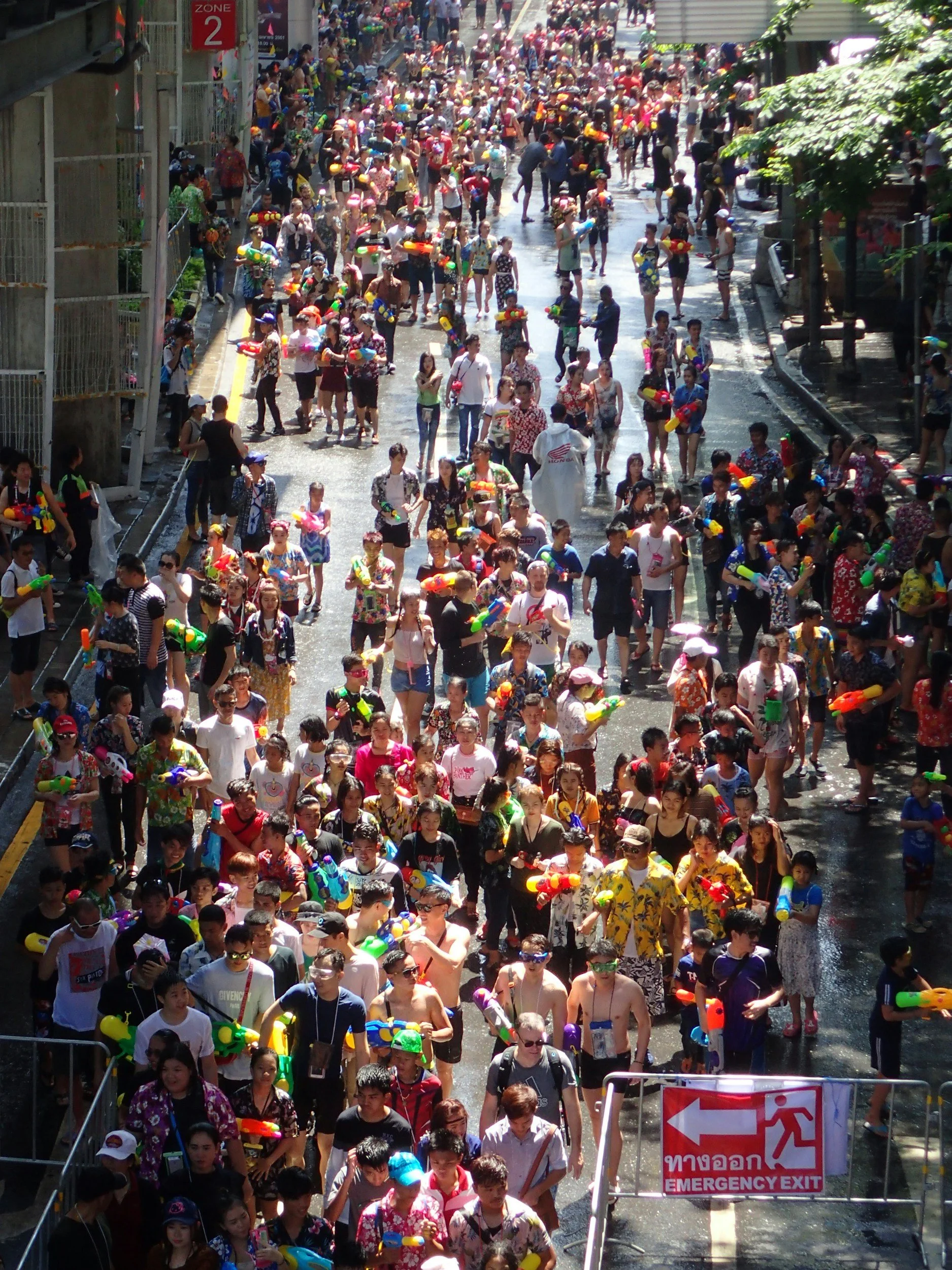 Crowd of people wearing summer clothes and holding water guns crossing a street on a sunny day, with an emergency exit sign visible in the foreground.