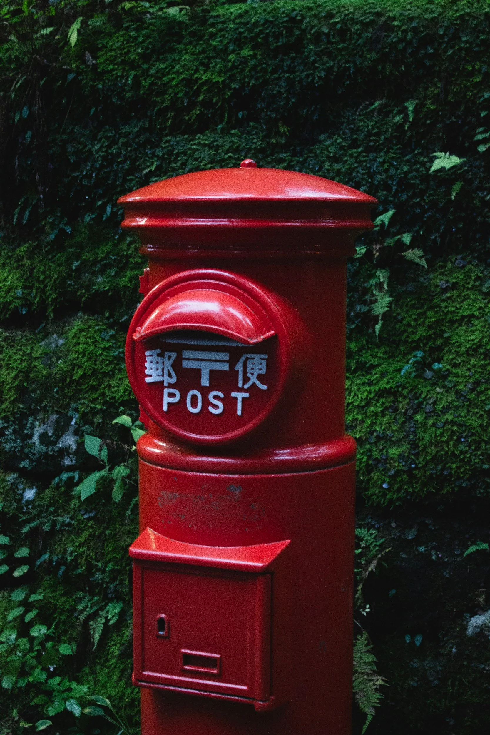 Red mailbox with Japanese characters on the front, surrounded by green foliage.
