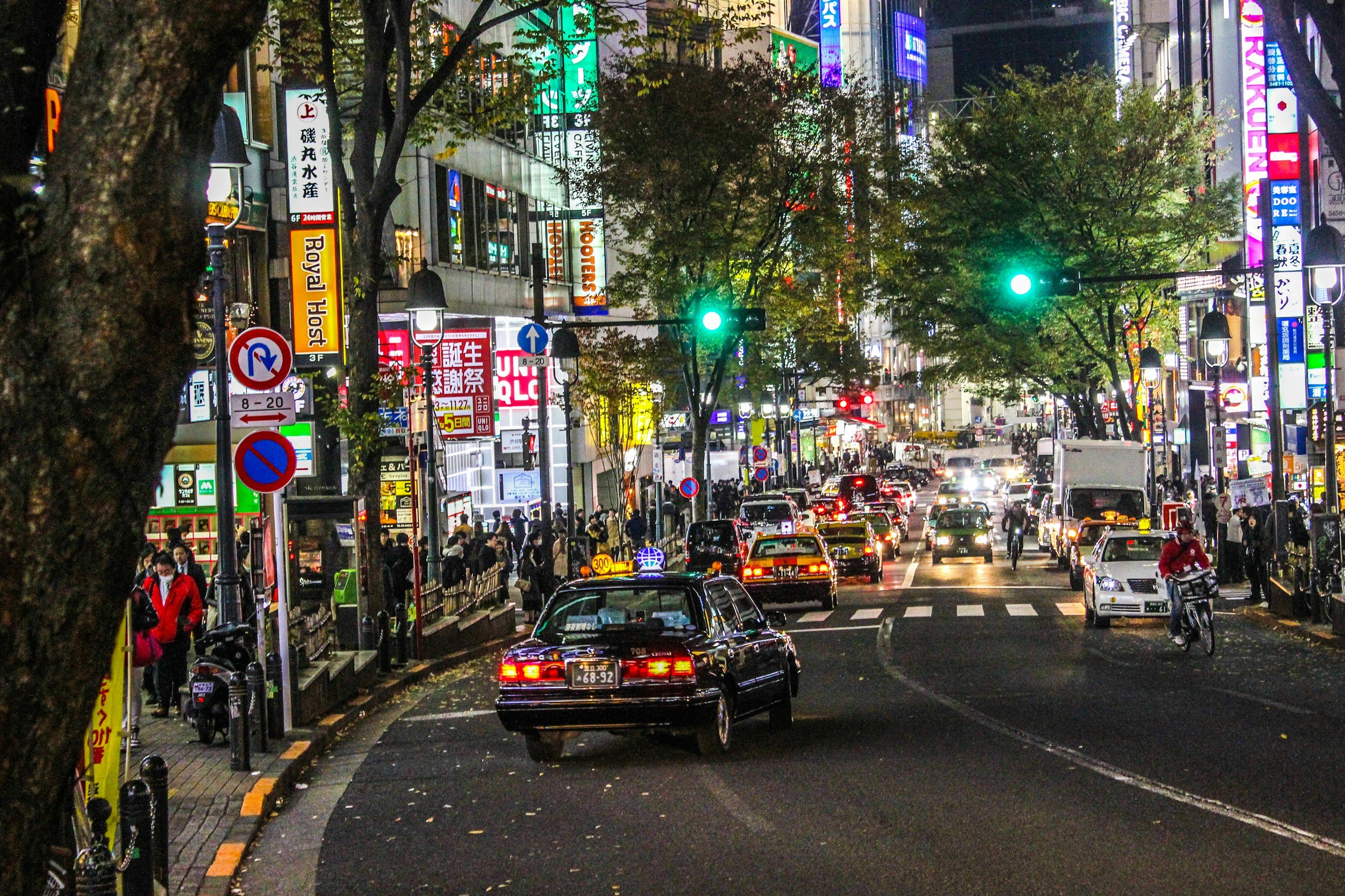 A busy city street at night illuminated by numerous colorful neon and digital billboards. Traffic, including cars and a motorcycle, is moving through the intersection, and pedestrians are walking along the sidewalks. Trees line the street, and various signs and storefronts are visible.