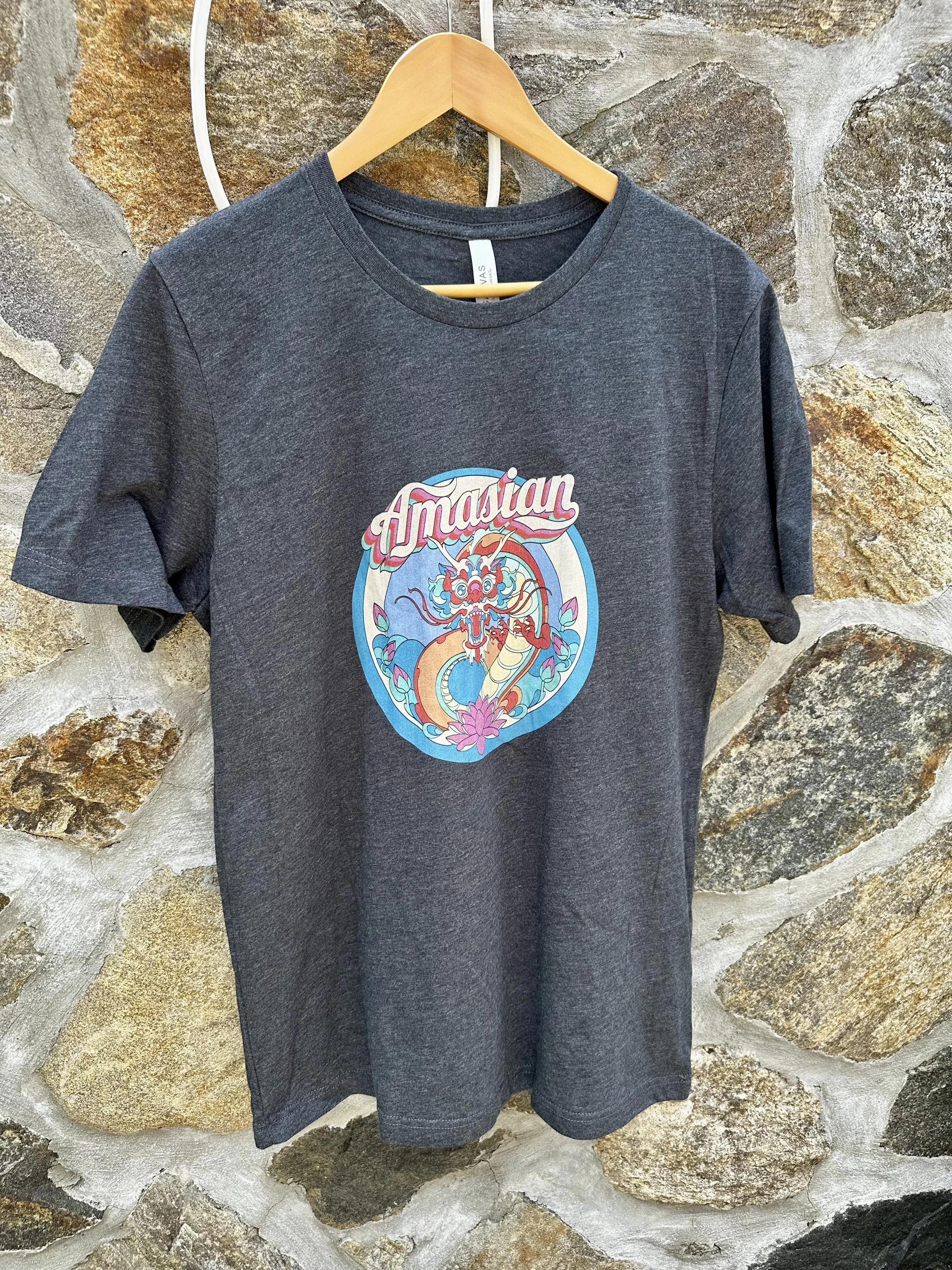 A gray T-shirt with the Amasian original graphic dragon logo graphic design with the word 'Amasian' on a hanger against a stone wall.