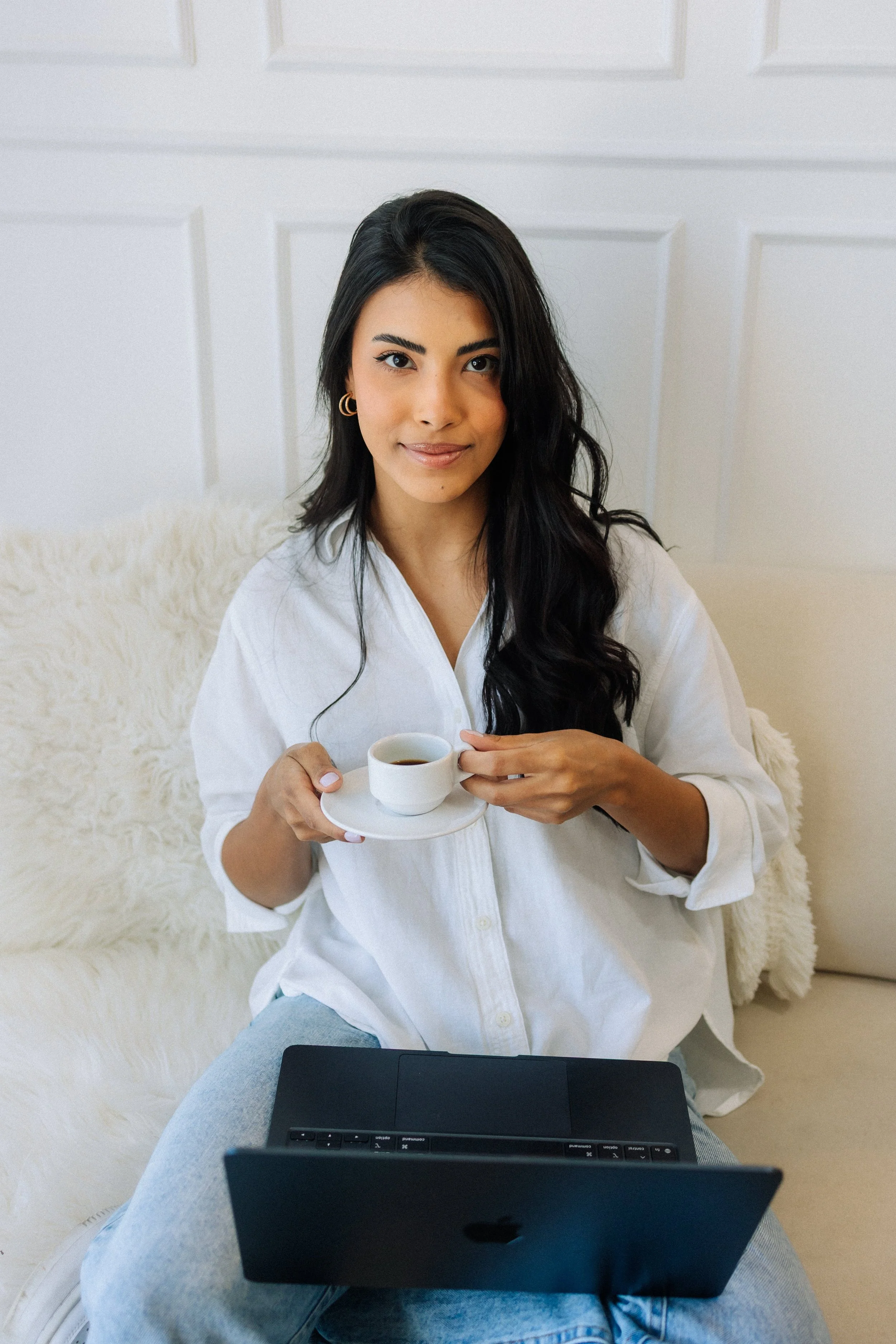 A woman with dark hair and earrings sitting on a fluffy white surface, holding a cup of coffee, looking at the camera, with a laptop on her lap.