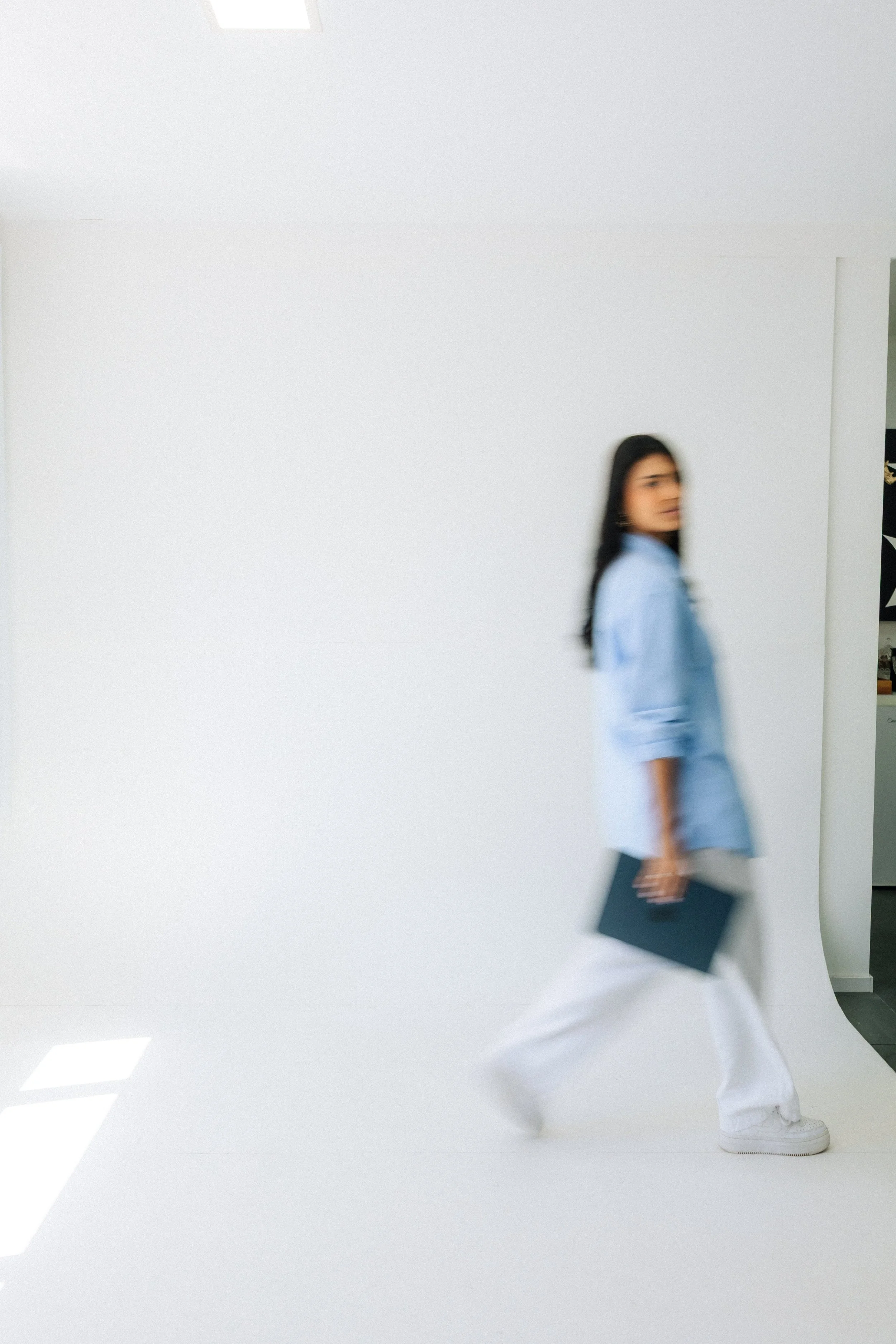 A woman with dark hair, wearing a blue jacket and white pants, walking and holding a black folder, in front of a plain white wall with bright light coming from above.