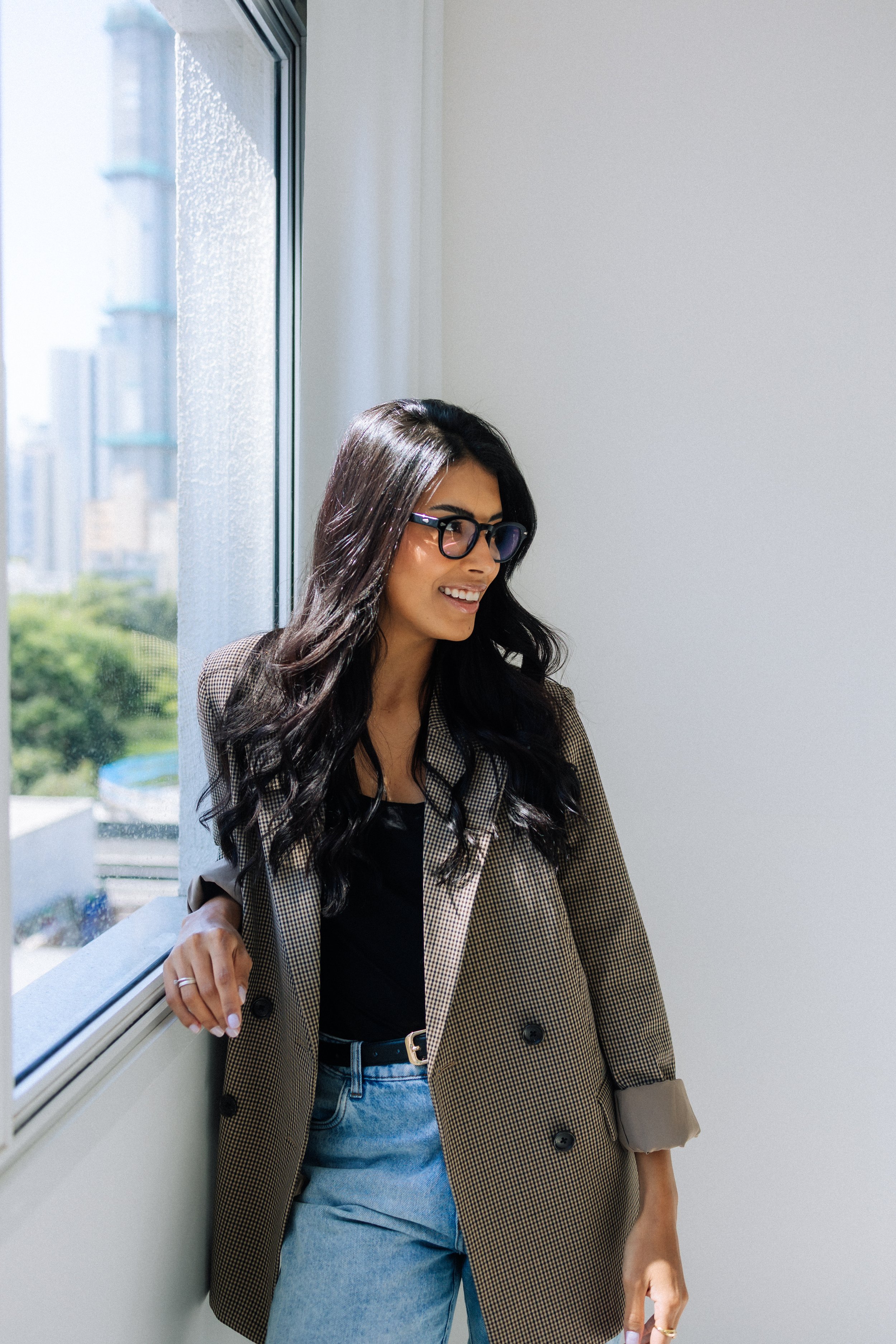 A woman with long dark wavy hair and glasses smiling while sitting by a window, wearing a plaid blazer, black top, and high-waisted jeans.