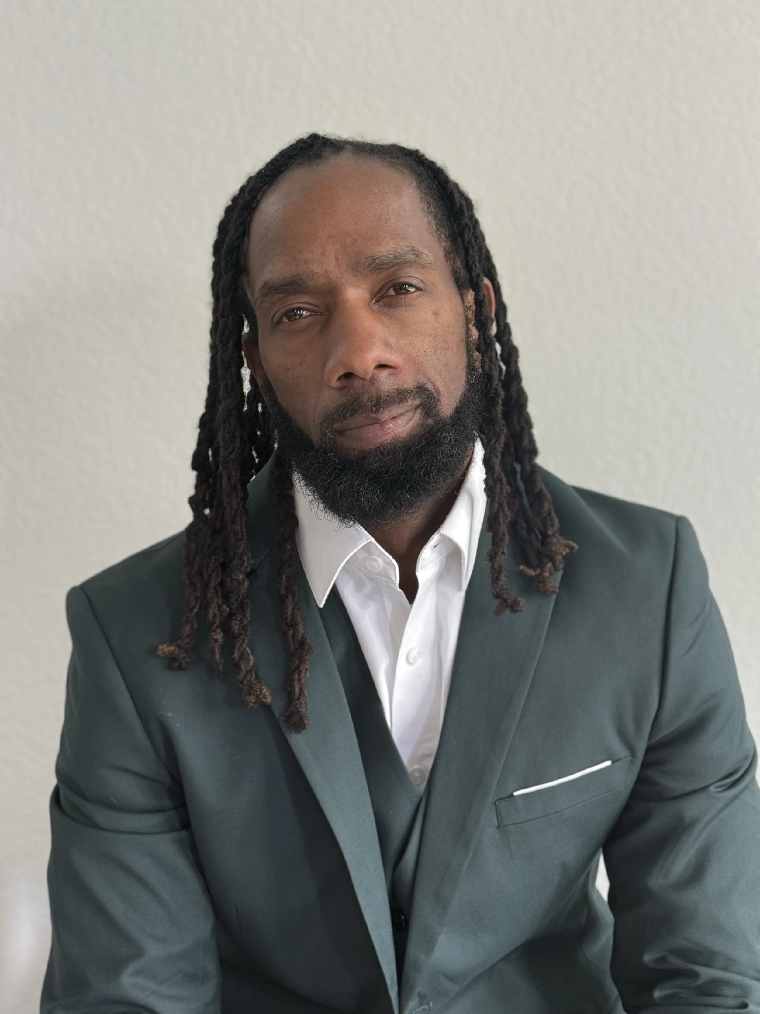 Michael Sykes dressed in a dark green suit and white shirt, with long dreadlocks and a beard, standing against a plain light-colored wall.