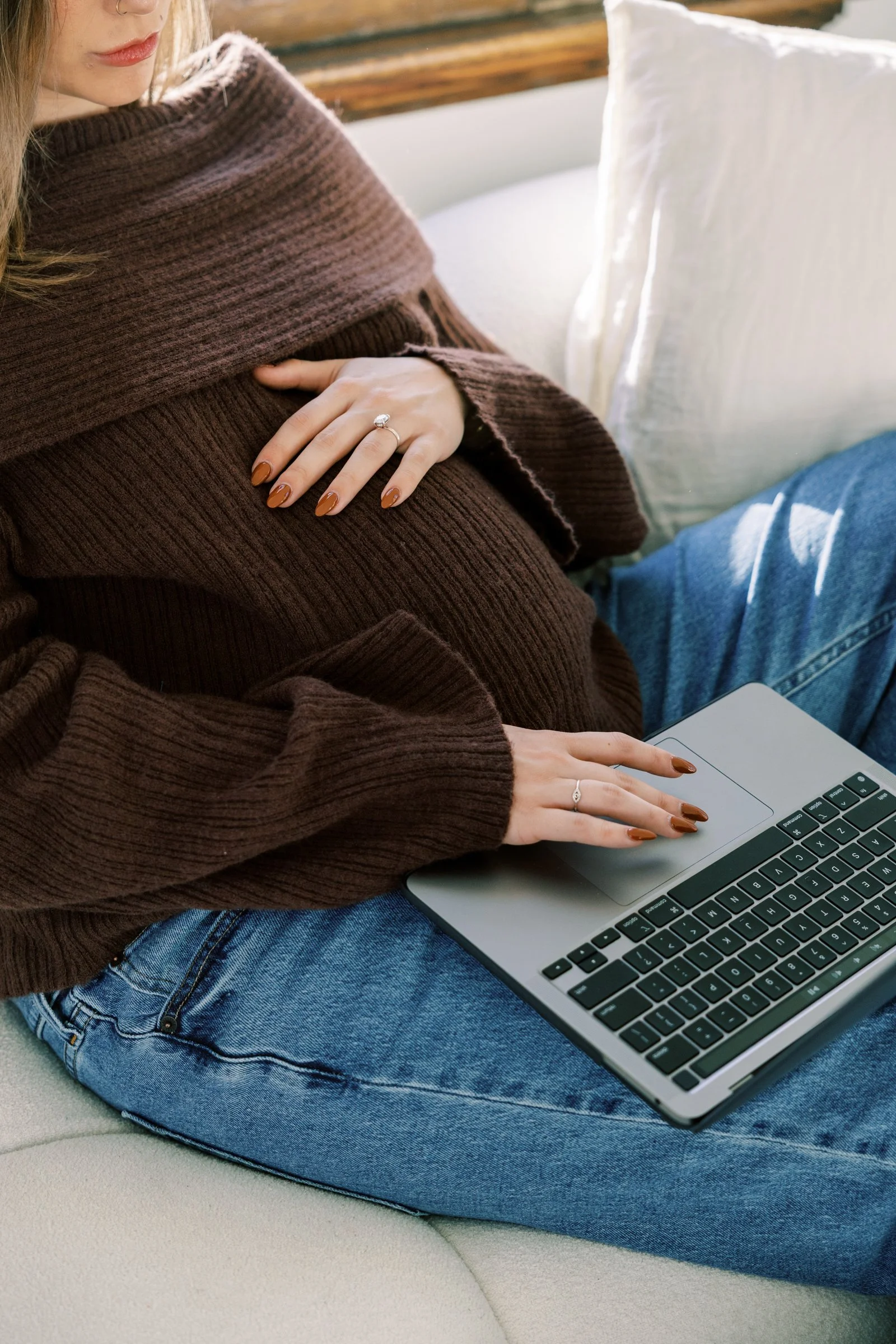 A woman sitting on a white couch using a silver laptop, wearing a brown sweater and blue jeans, with her left hand on the laptop keyboard and her right hand on her abdomen. She has manicured brown nails and is wearing rings.