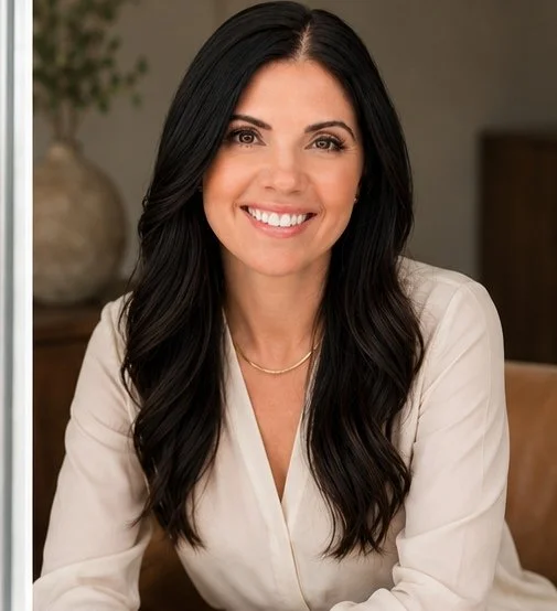 A woman with long dark hair and a white blouse smiling at the camera indoors.