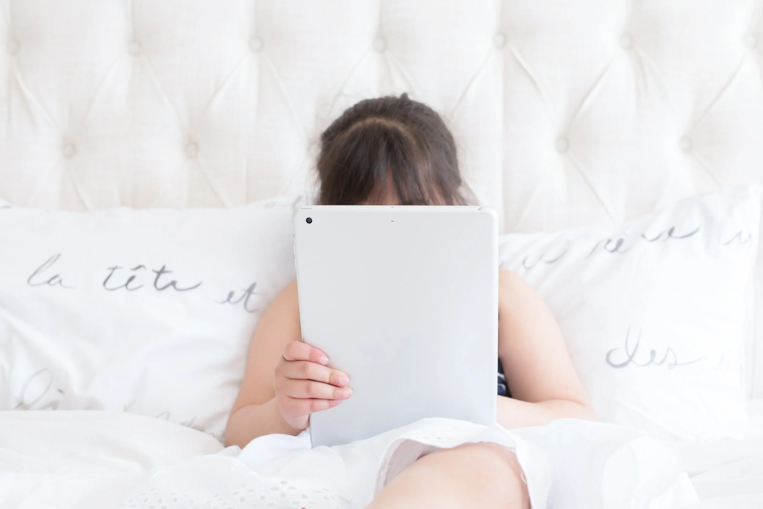 A young child sitting on a bed, holding a tablet in front of their face and looking at it. The bed has white bedding with cursive writing, and the headboard is quilted and padded.