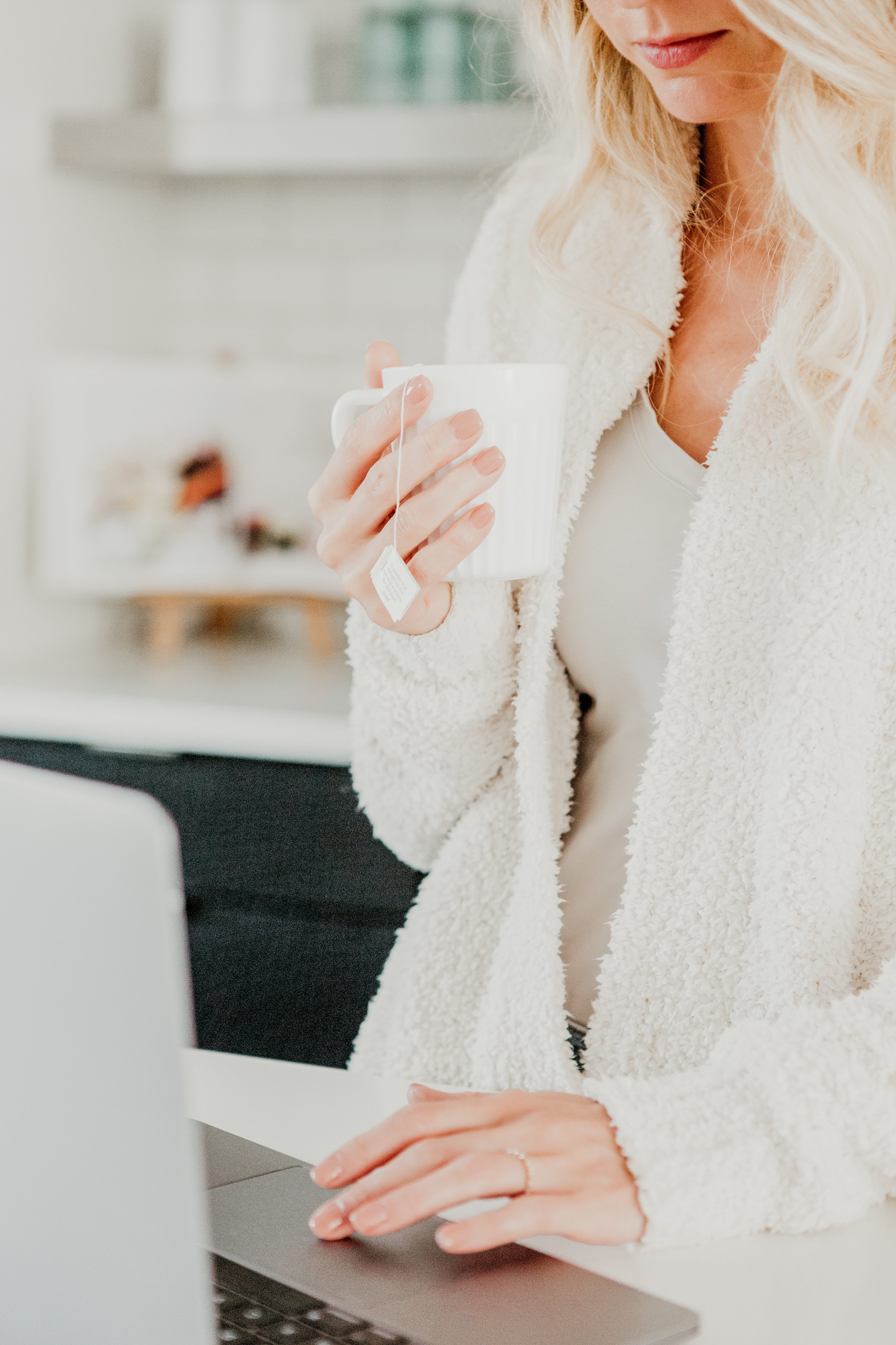 A woman wearing a white fluffy jacket holding a white mug, sitting at a desk with a laptop, in a bright room.
