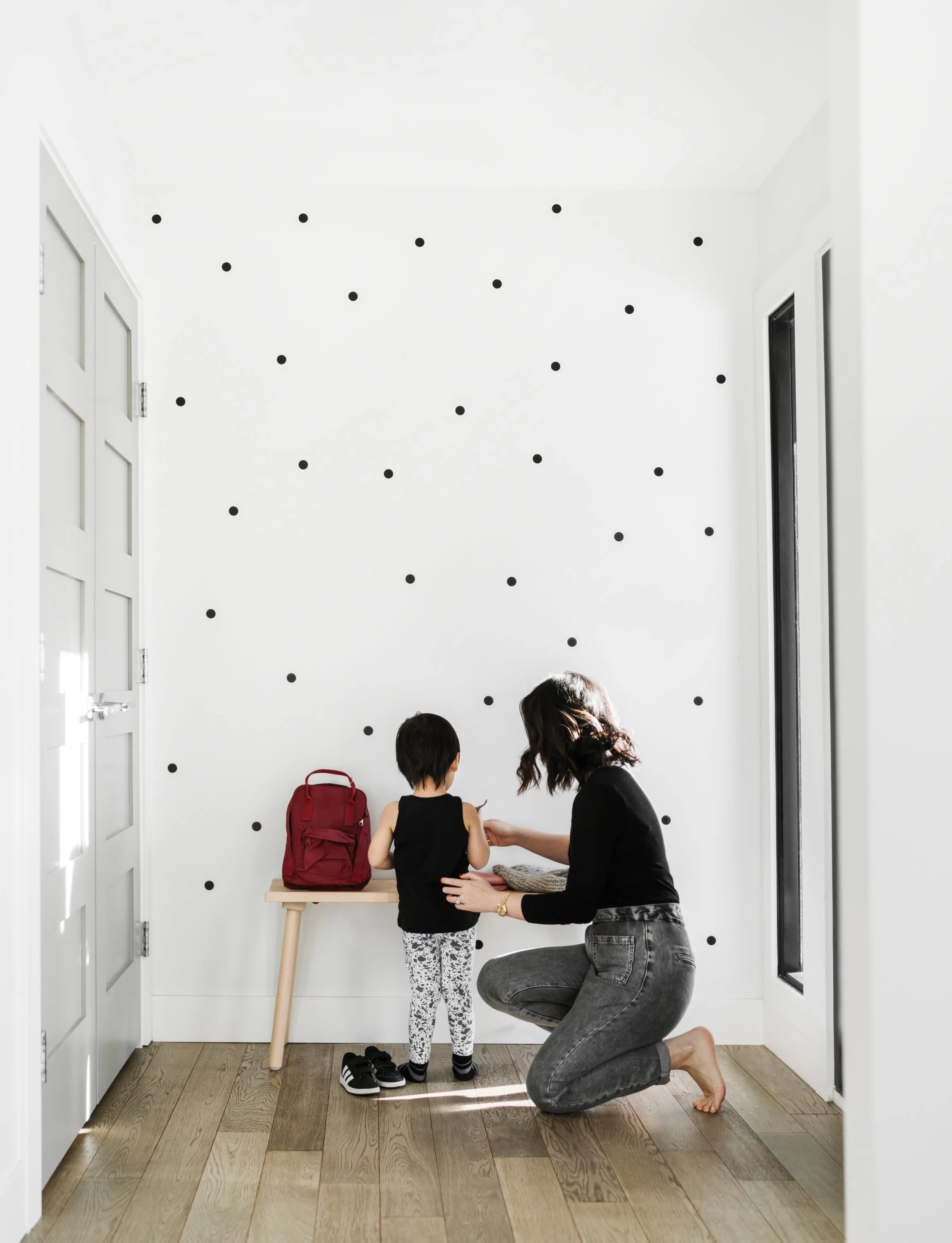 A woman is kneeling on the wooden floor in a white room with a black dotted wall, helping a young girl tie her shoes. The girl stands with her back to the woman, wearing leopard-print leggings and a black top. There is a red backpack on a table beside them.