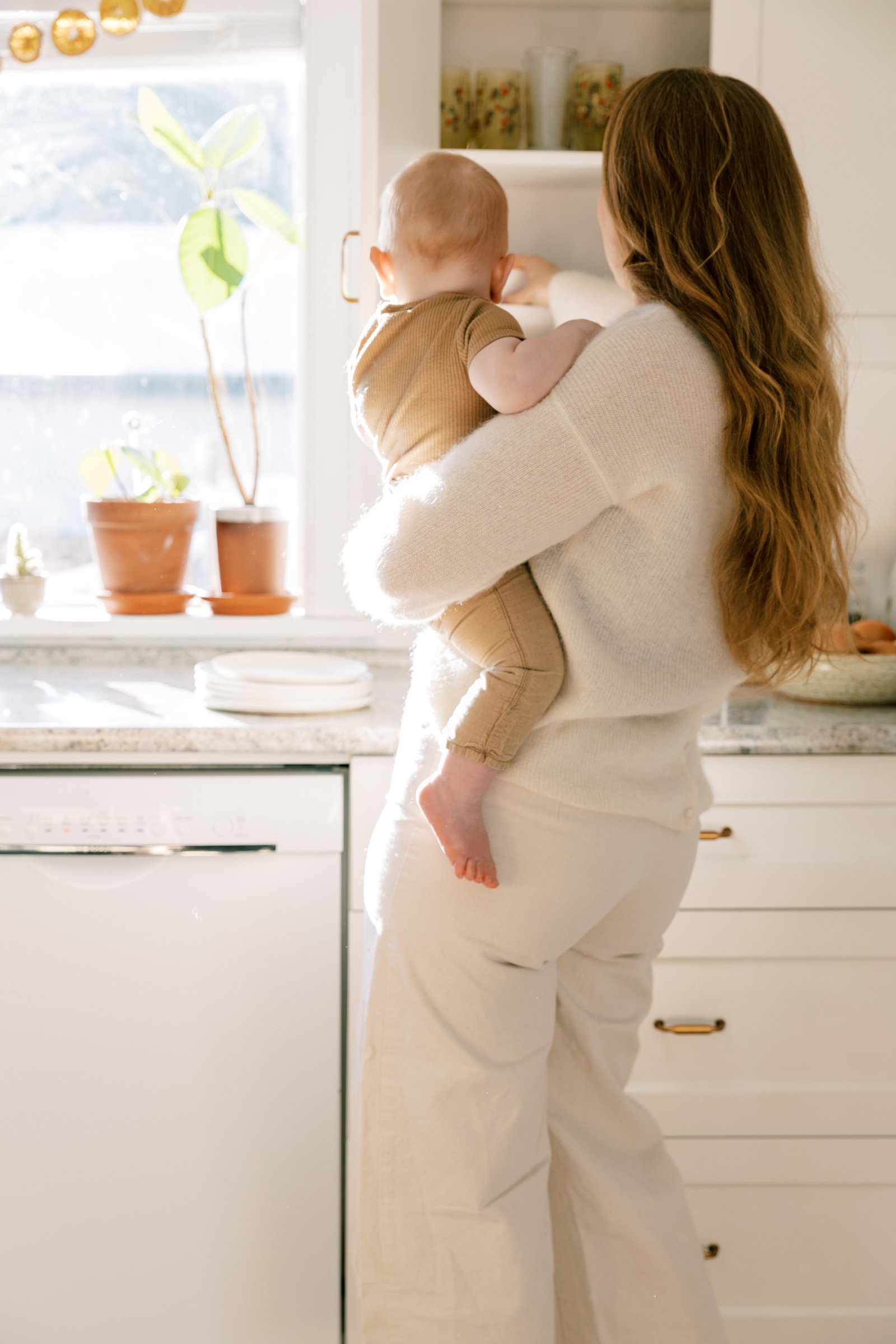 A woman with long red hair holding a young child while standing in a bright kitchen near a white cabinet. The child is reaching into the cabinet. Sunlight streams through the window, illuminating potted plants and kitchen counters.