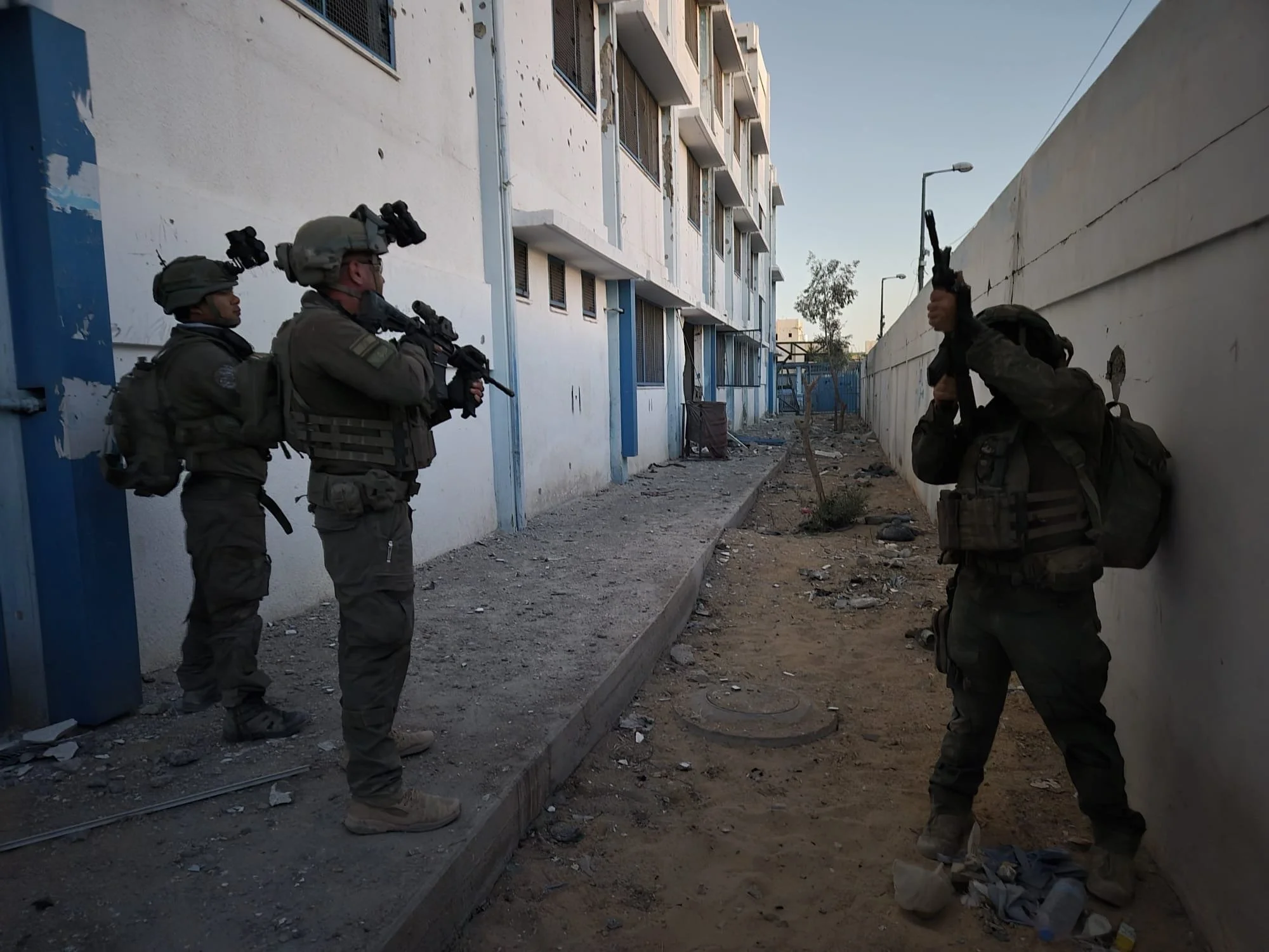 Four IDF soldiers in combat gear, wearing helmets with night vision mounts, standing in a narrow alleyway with dilapidated buildings on one side and a high wall on the other. Documentary footage from The Prophet Documentary by Melissa Francis.