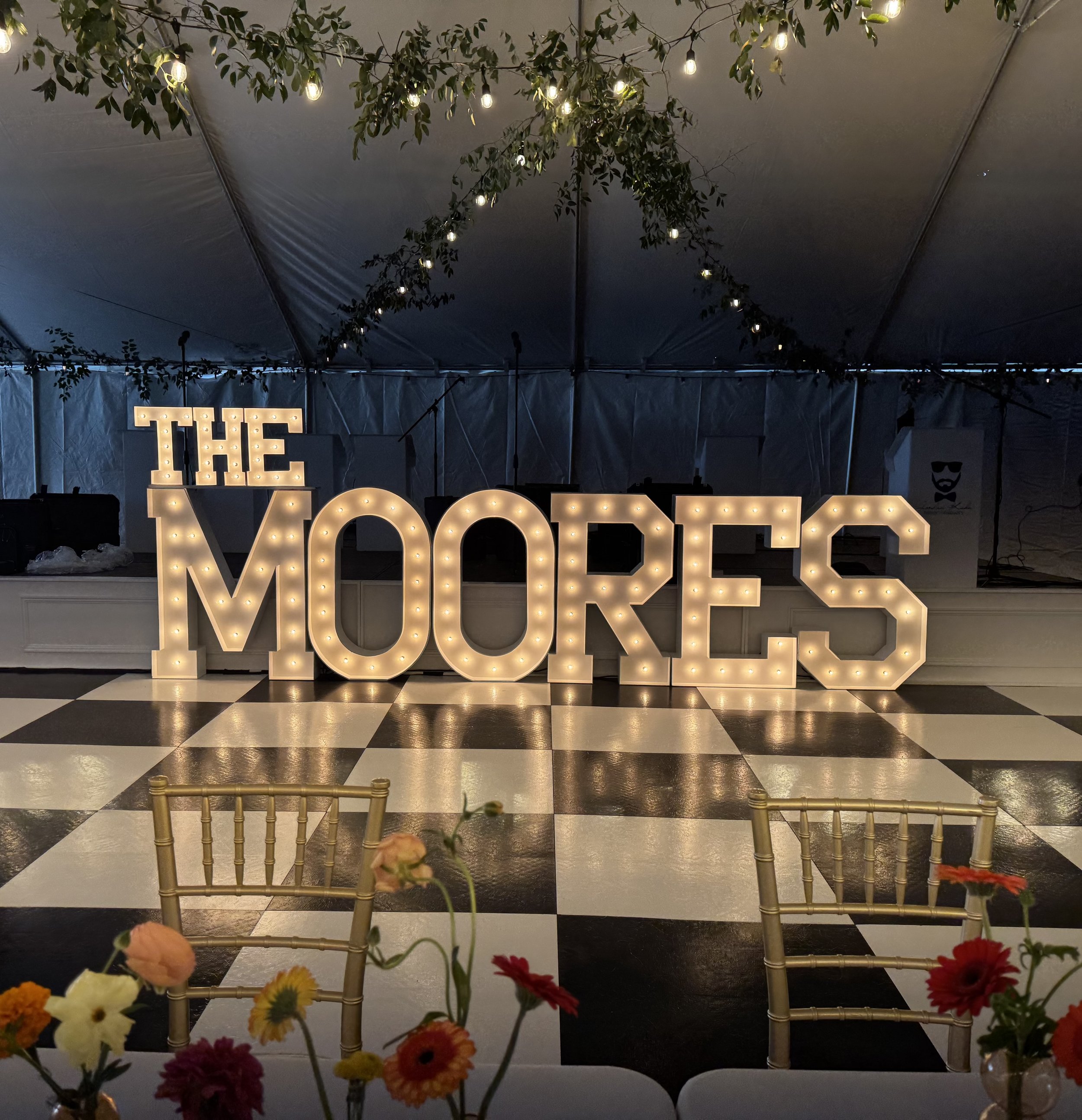 Decorative indoor area with hanging string lights, green foliage, and illuminated marquee letters spelling "THE MOORES" on a stage with a black and white checkered floor, surrounded by chairs and flowers.