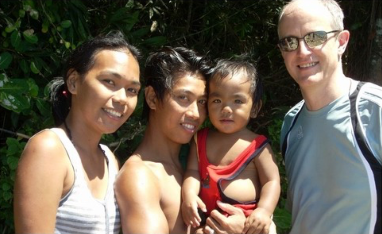 Group of four people, two women, a man, and a young child, smiling outdoors with green foliage in the background.