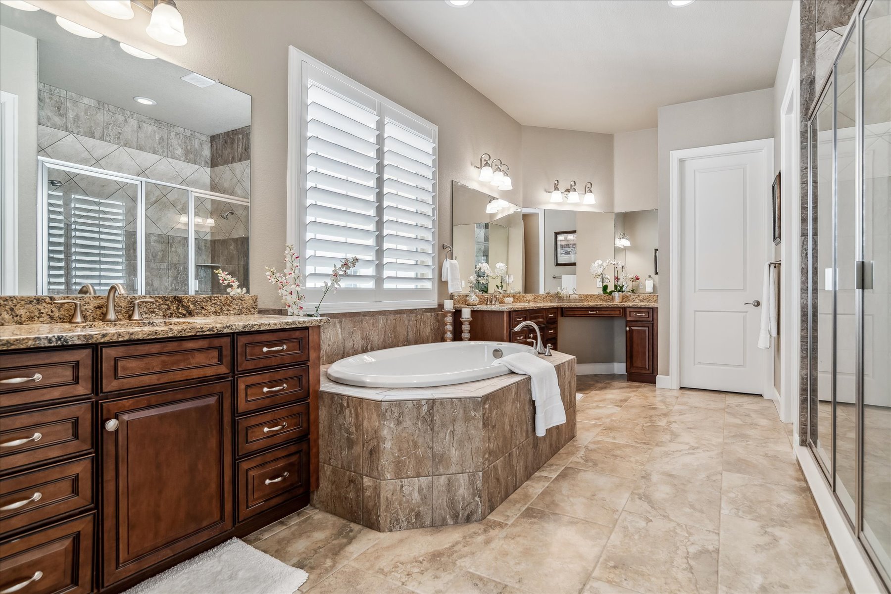 Bathroom with a double vanity with dark wood cabinets and granite countertops, a large bathtub with a tiled surround, a glass-enclosed shower, and a window with white plantation shutters. The room has beige walls and tiled floors.