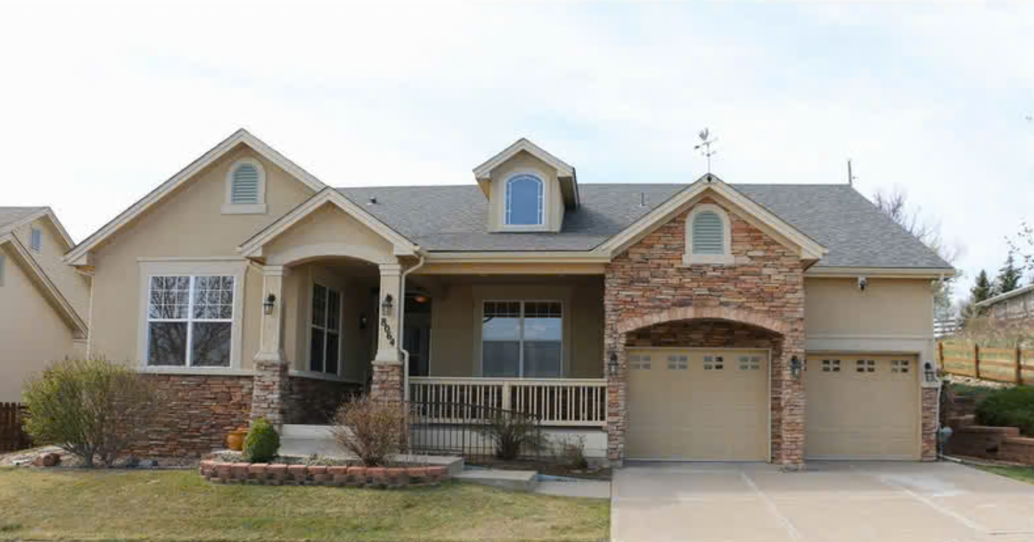 A two-story house with beige walls, brick accents, and a gray roof. It has a front porch with columns, a double garage, and a small front yard with bushes and a lawn.