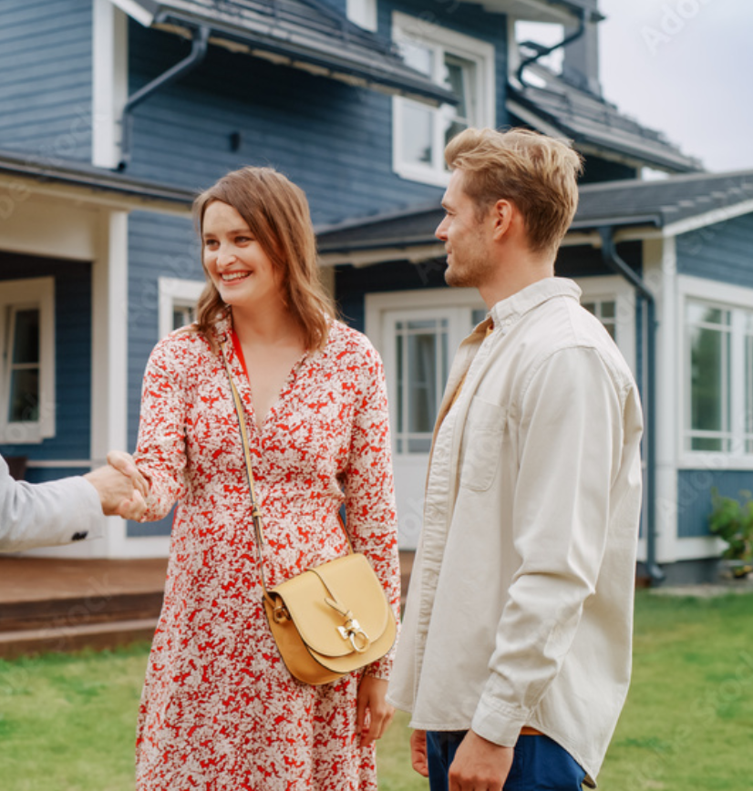 A young woman in a red and white floral dress is shaking hands with a person out of frame, while a young man in a cream jacket and blue pants looks on, standing outside a blue house with white trim.