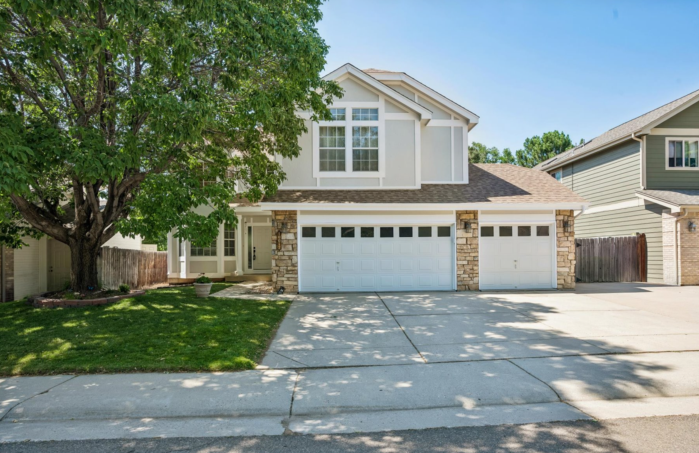 A two-story house with a white exterior, stone accents, and a garage. There is a large green tree in the front yard, partially blocking the view of the house. The driveway and sidewalk are visible.