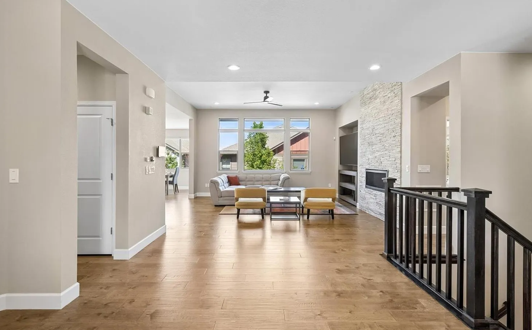 Open concept living room with wooden flooring, a white tufted sofa, a coffee table, yellow chairs, large windows showing trees outside, a white brick fireplace, and a ceiling fan.