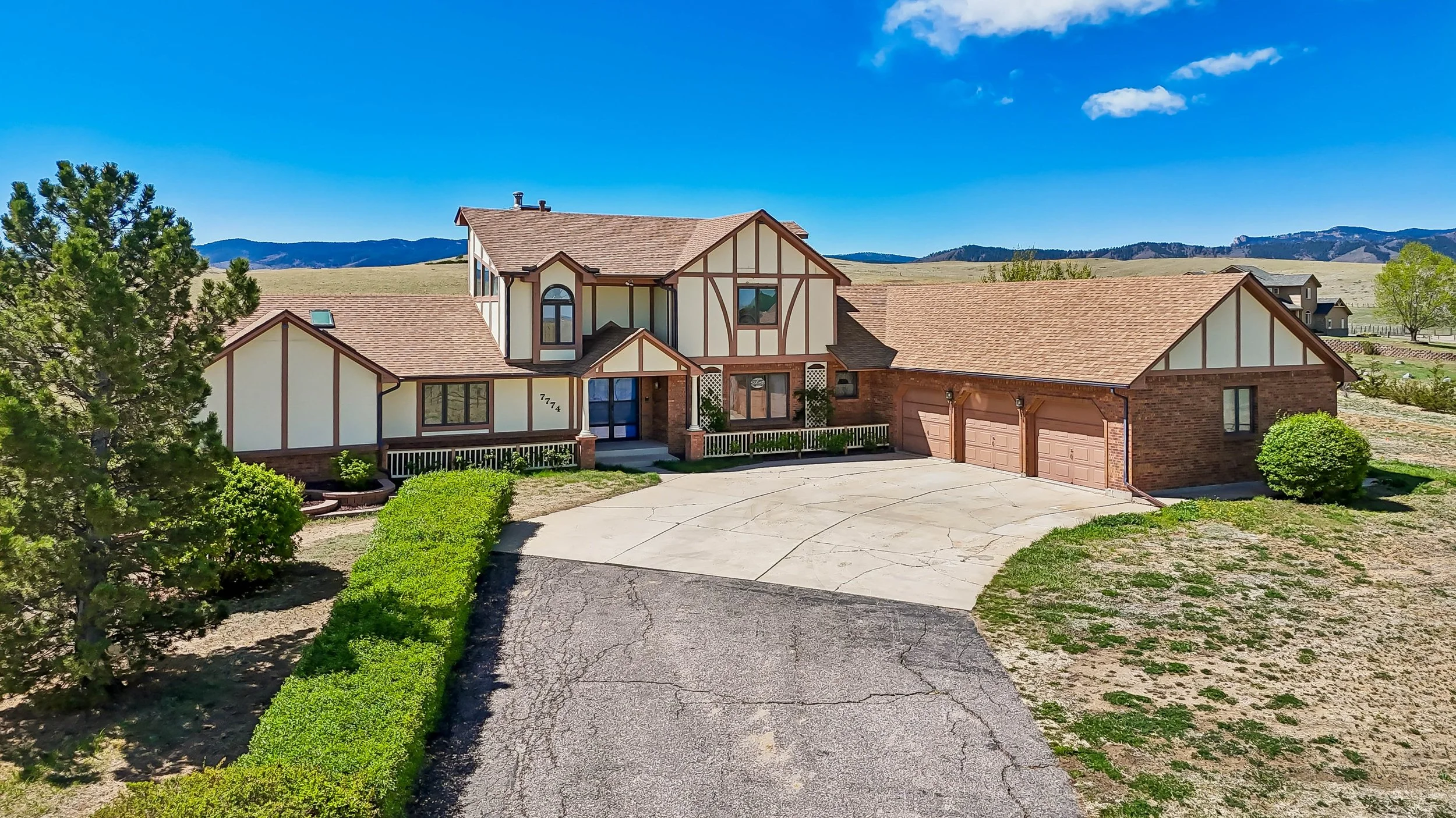 A large house with a mix of brick and Tudor-style exterior, brown shingle roof, attached three-car garage, front yard with green bushes, a driveway, and a surrounding landscape with mountains in the background under a clear blue sky.