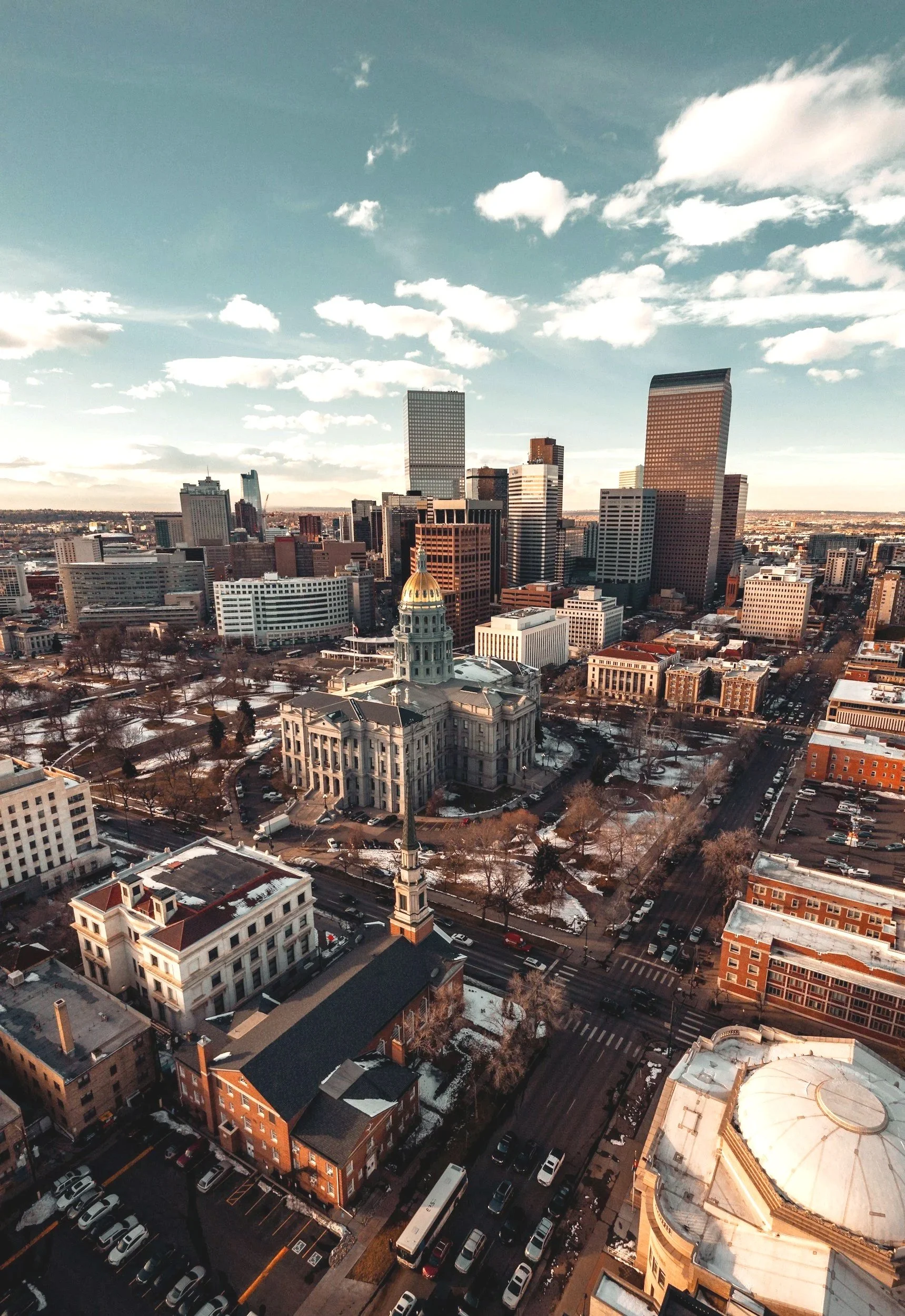 A cityscape with a mix of modern high-rise buildings and historic structures, showing snow on the ground and a cloudy sky during sunset.
