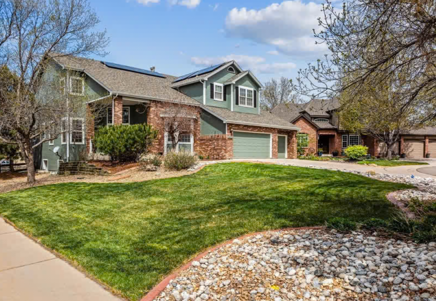 Front view of a modern two-story house with a landscaped yard, including a well-maintained lawn, stone and brick exterior, and surrounding trees under a partly cloudy sky.