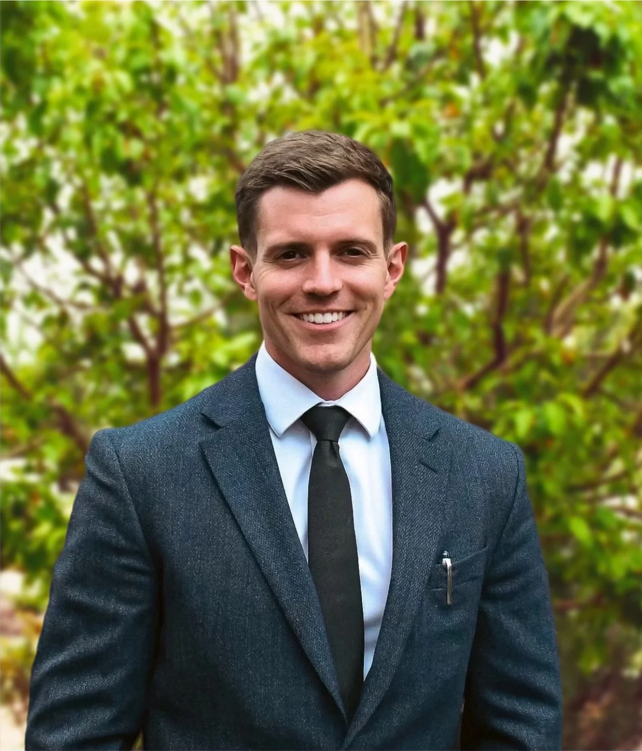A man in a dark suit with a white shirt and black tie smiling outdoors with green leaves in the background.