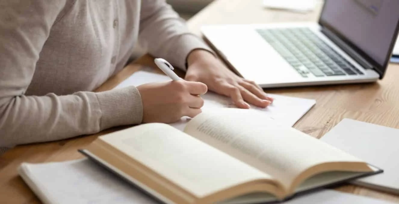 Person writing in a notebook with a laptop open nearby on a wooden desk.