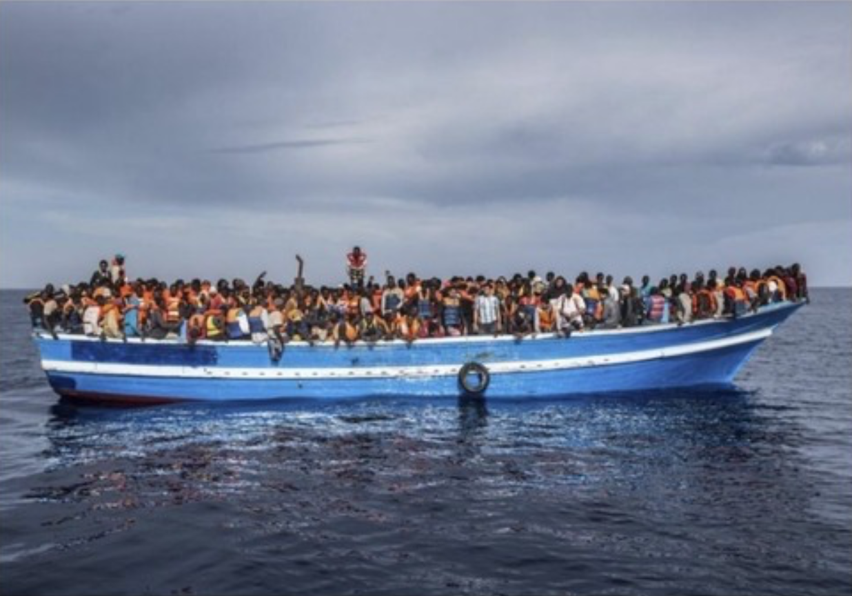 Large boat filled with many people wearing life jackets, sailing on the ocean under a cloudy sky.