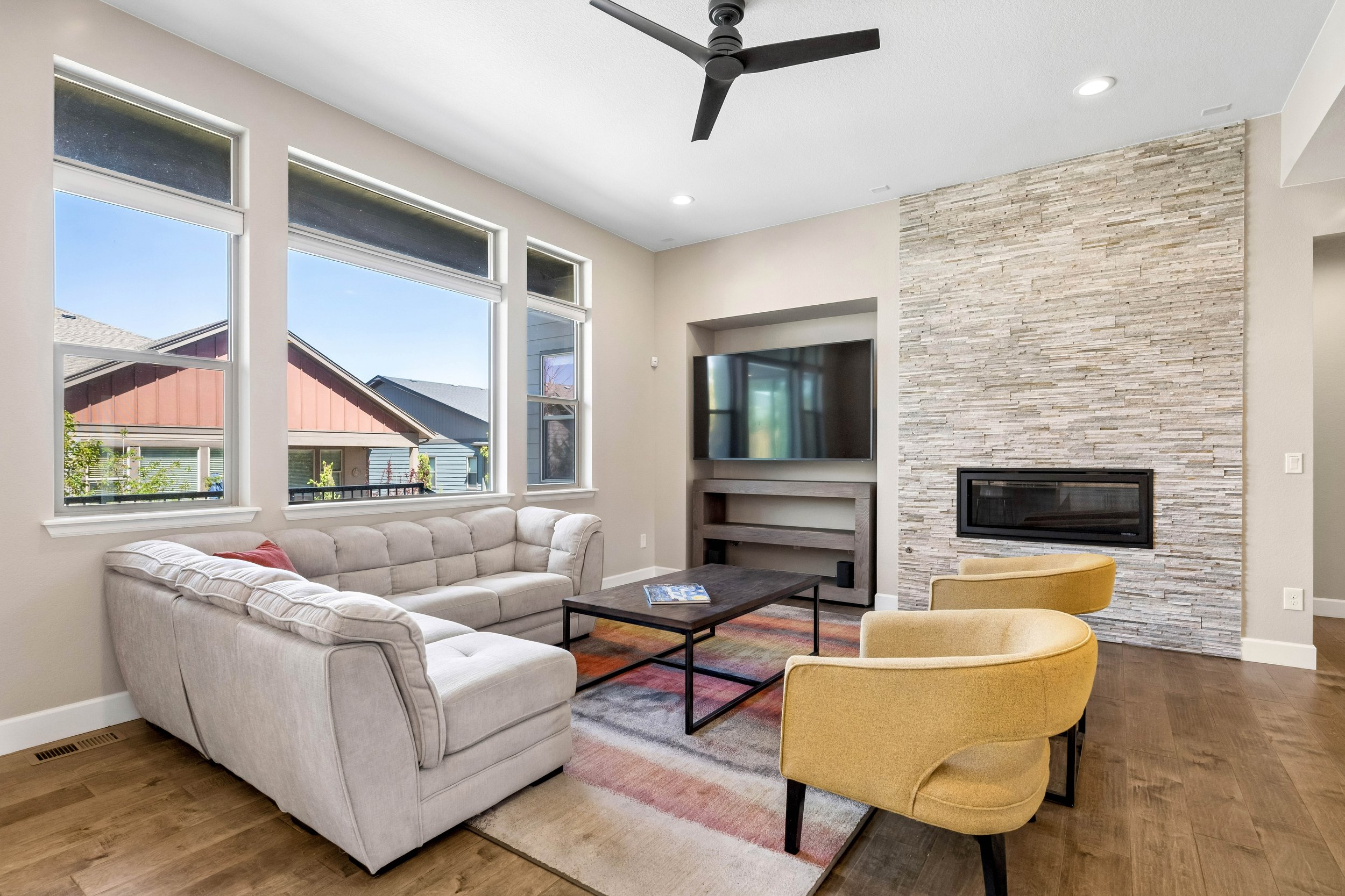 A modern living room with large windows, a beige sofa, a coffee table, a media console, a flat-screen TV, a stone accent wall around a fireplace, and a ceiling fan.