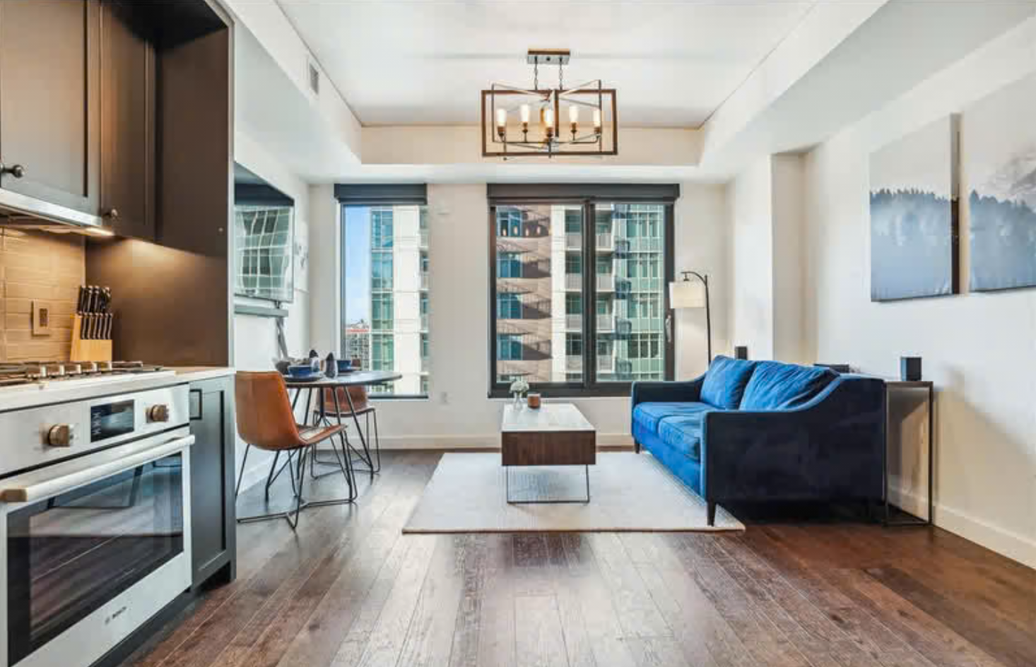 Living room with large windows, blue sofa, wooden coffee table, art on white wall, dining area with two chairs and table, kitchen with stove and knife set, hardwood floors, and modern lighting fixture.