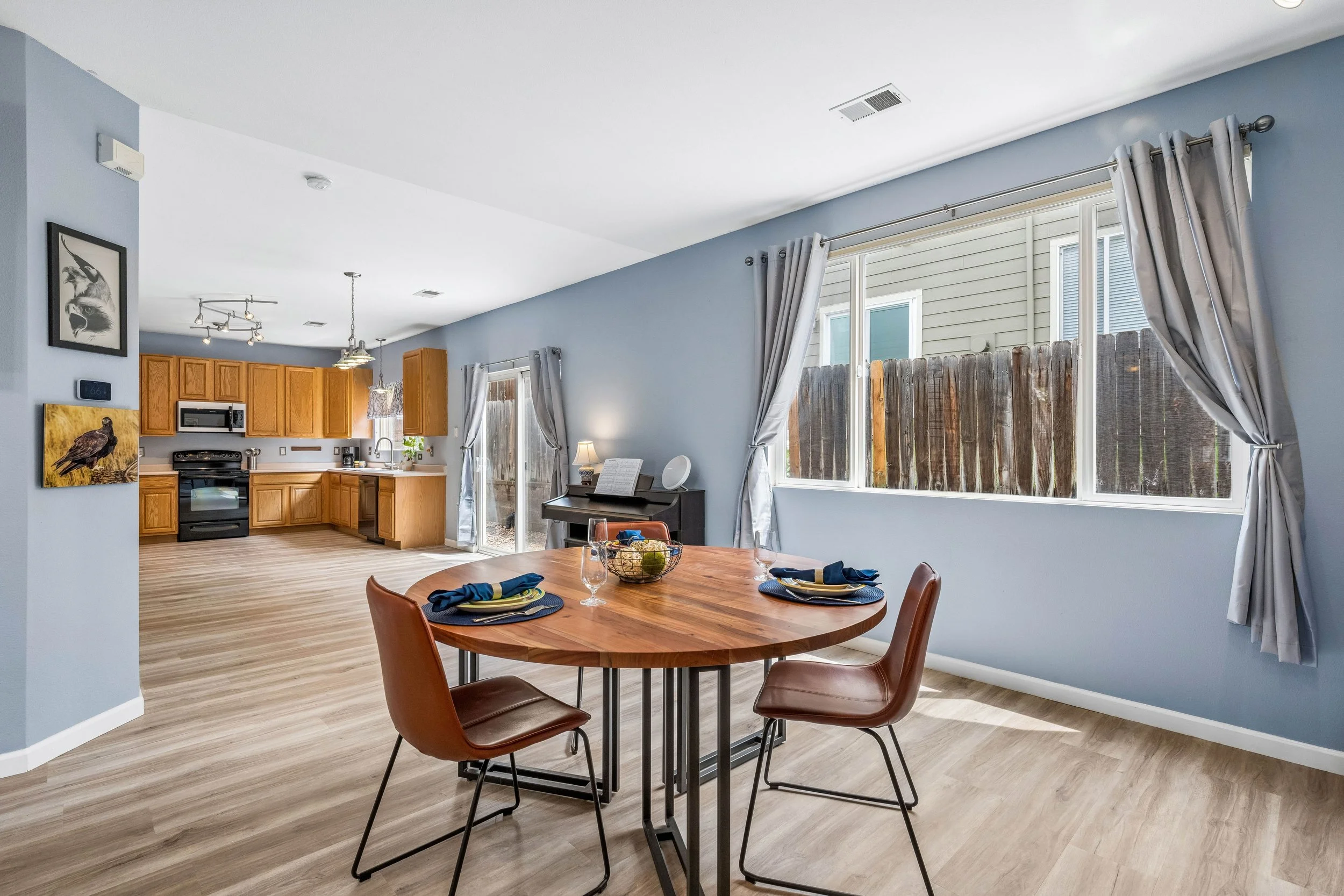Interior of a modern kitchen and dining area with light blue walls, wooden cabinets, a round wooden table set for four, and large windows with light-colored curtains.