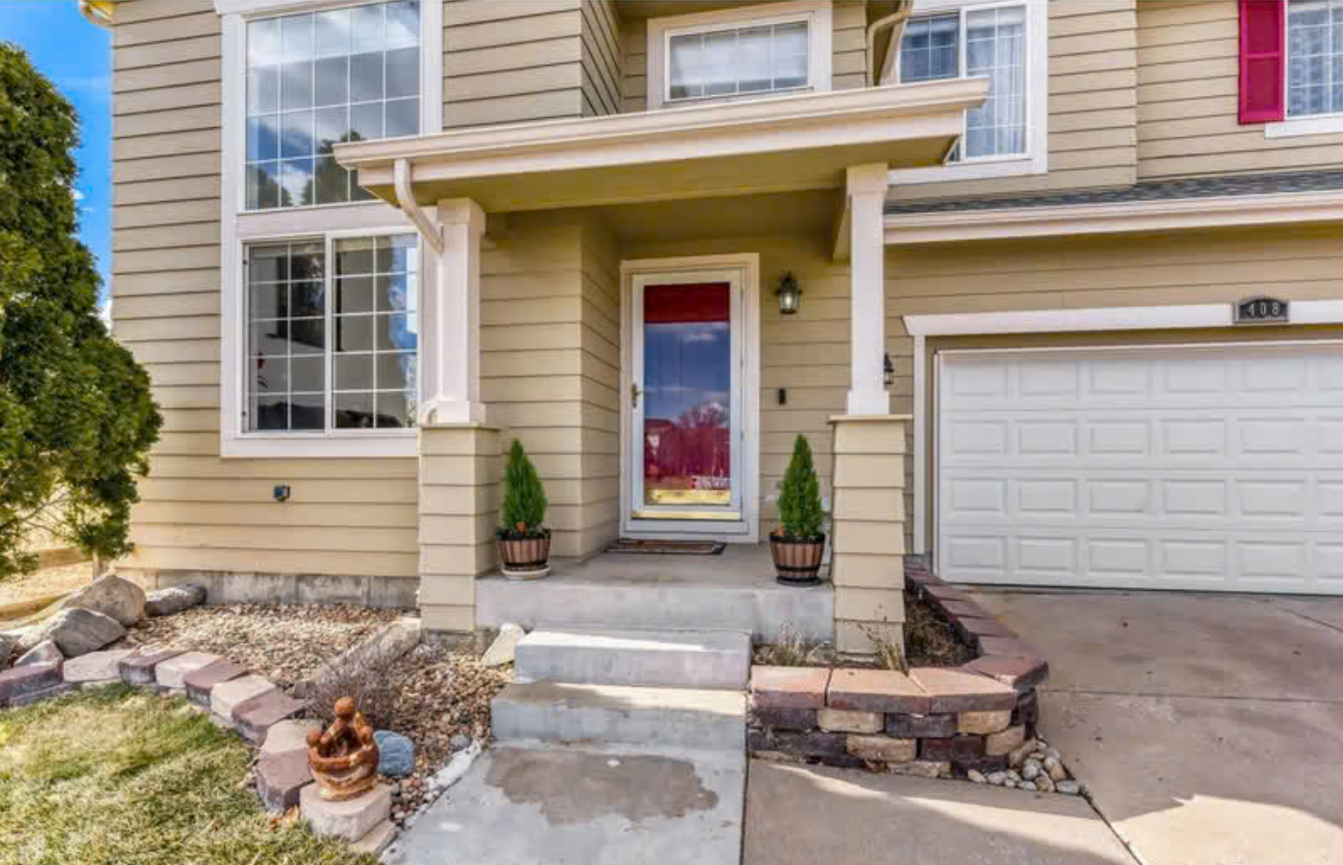 Front view of a beige two-story house with a small front porch, red glass door, potted plants, large window on the left, white garage door, and concrete driveway under partly cloudy sky.