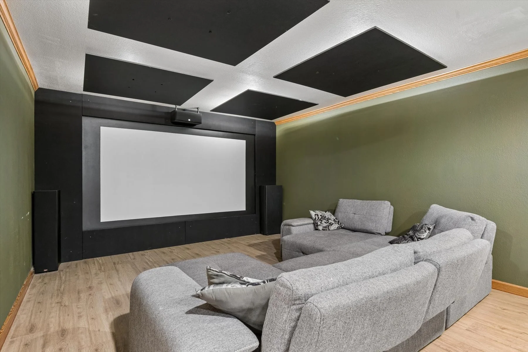 A home theater room with a large white projection screen surrounded by black and green walls. Ceiling has black acoustic panels. There is a gray sectional sofa with decorative pillows, and wood flooring.