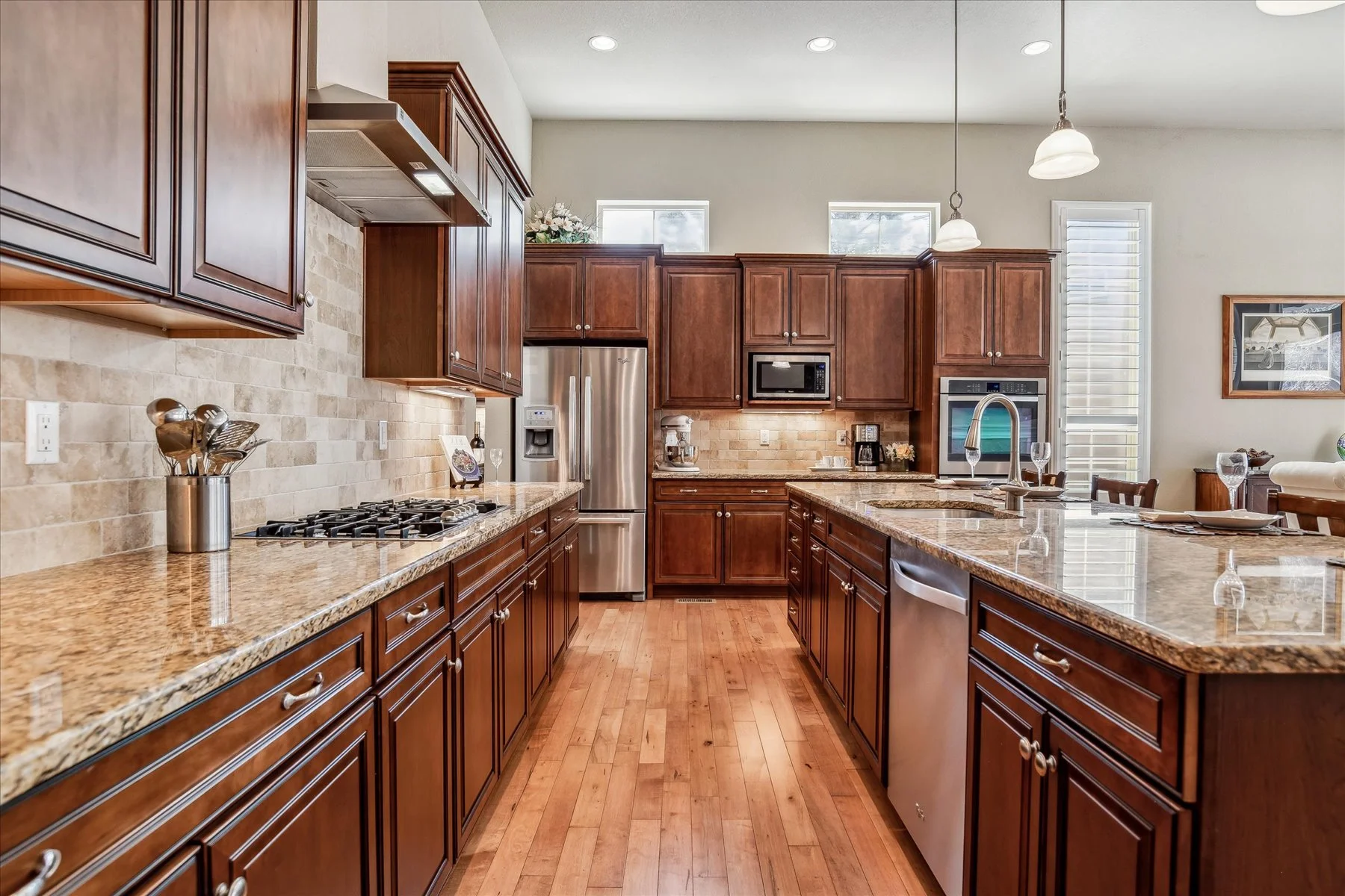 A spacious kitchen with wooden cabinets, granite countertops, stainless steel appliances, and hardwood floors. The kitchen features a large island with a sink and pendant lights.