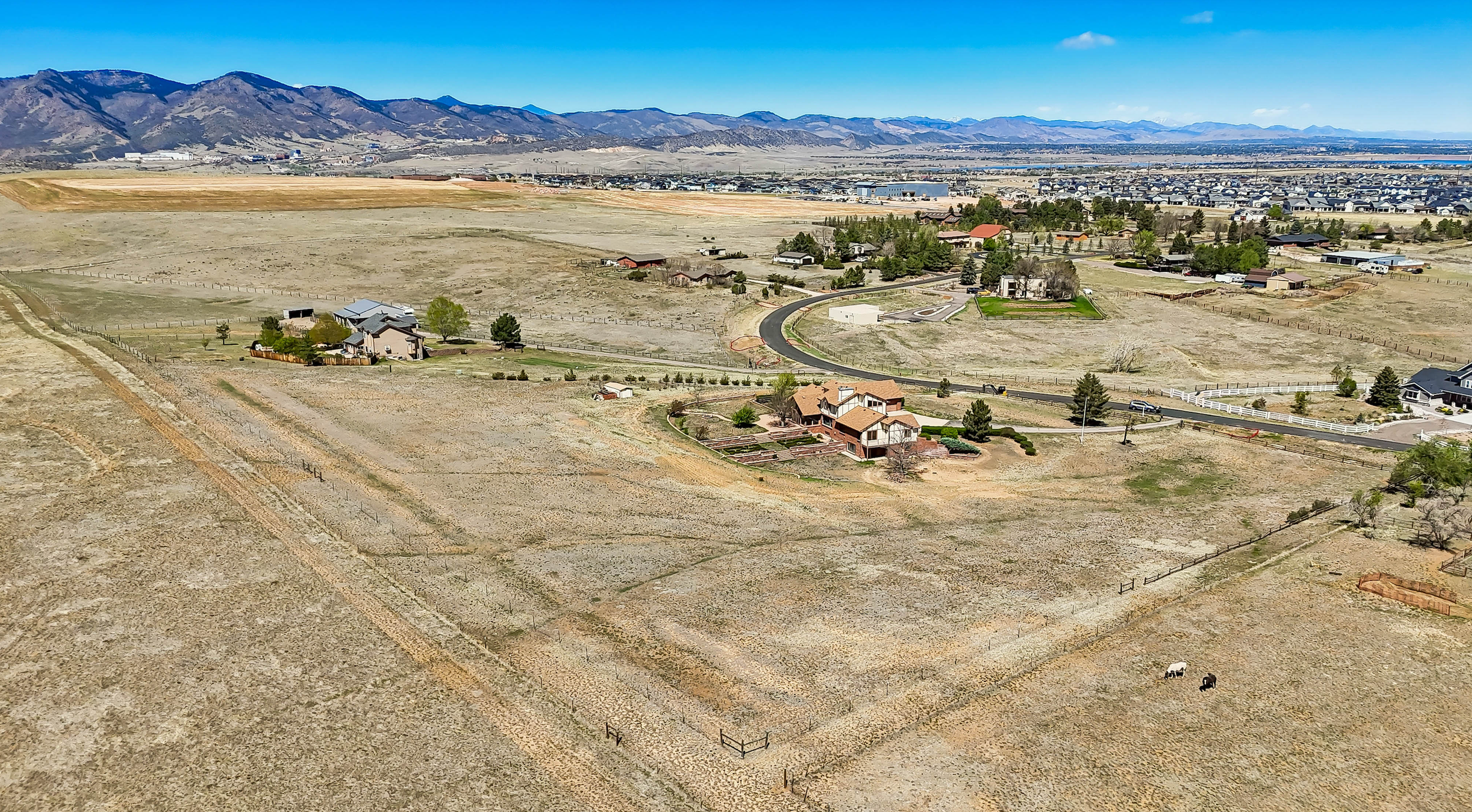 Aerial view of a semi-rural area with houses, fenced properties, and open fields, with mountains in the background under a clear blue sky.