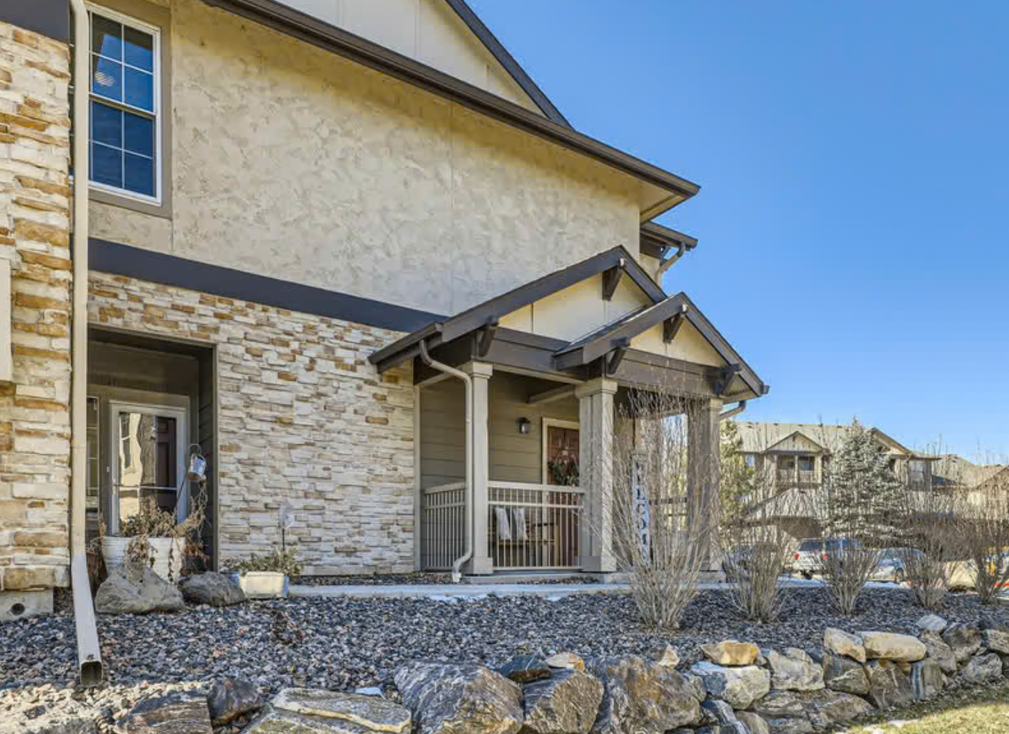 Front view of a modern house with a stone and stucco exterior, small porch with columns, and landscaped yard with rocks and shrubs.
