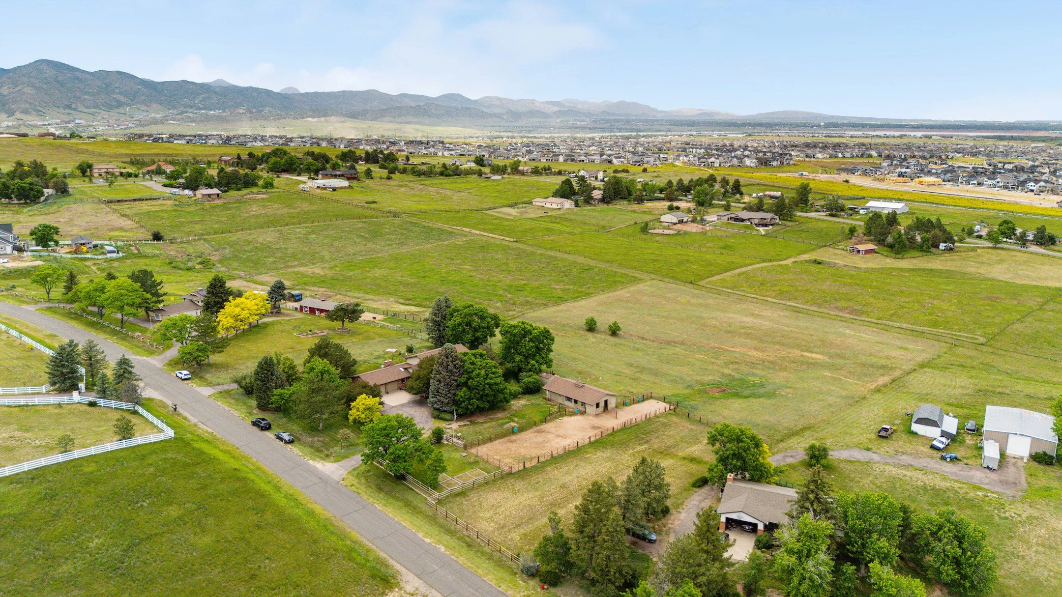 Aerial view of a suburban area with green fields, houses, and trees, with mountains in the background under a partly cloudy sky.