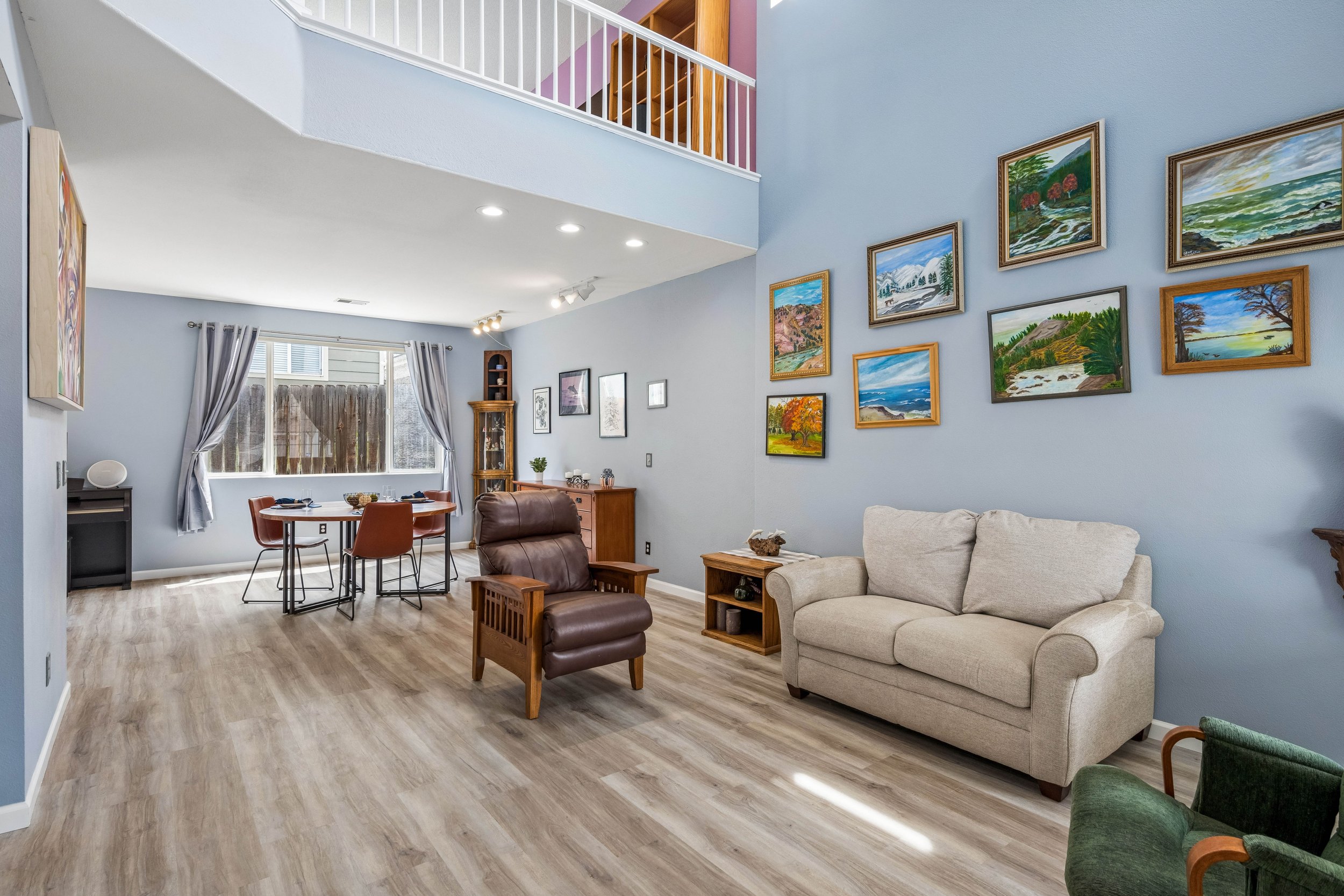 Open concept living and dining area with light blue walls, hardwood flooring, and a wall decorated with framed landscape paintings. The space includes a beige sofa, a green armchair, a wooden armchair, and a dining table with brown chairs. Large window with curtains provides natural light.