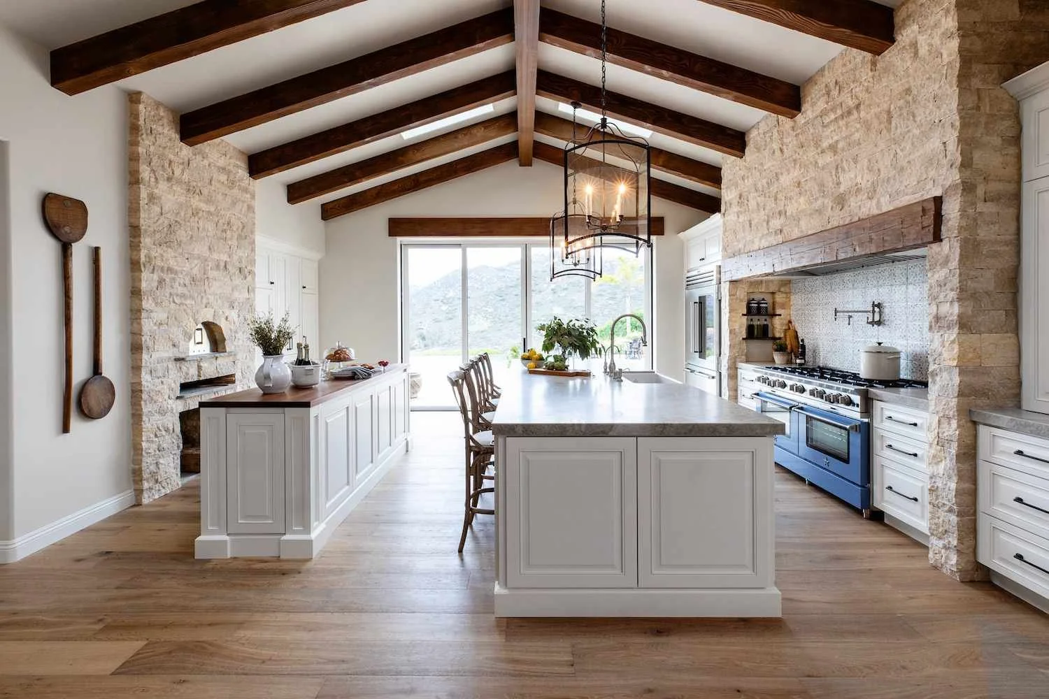 A spacious kitchen with white cabinets and a large island with seating. Exposed wooden beams on the ceiling, stone walls, and a hardwood floor. Large sliding glass doors at the back, natural light, and outdoor scenery visible.