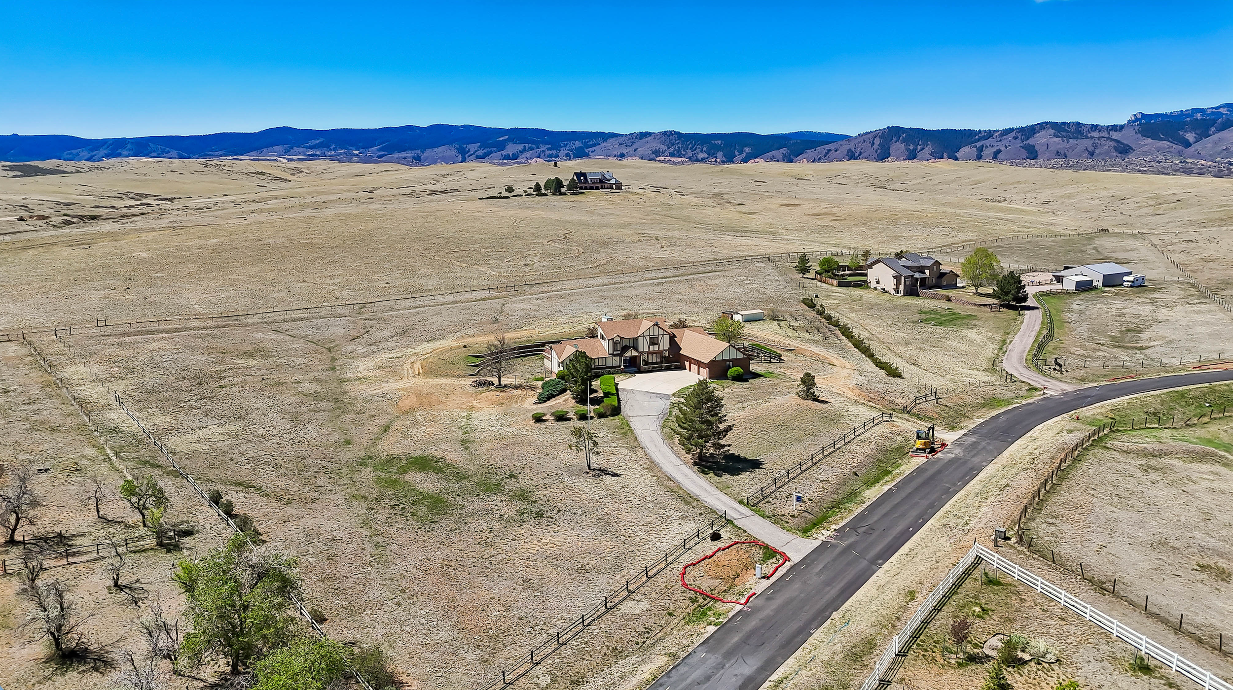 Aerial view of a rural neighborhood with houses, trees, fences, and a winding road, surrounded by open hills and mountains under a clear blue sky.